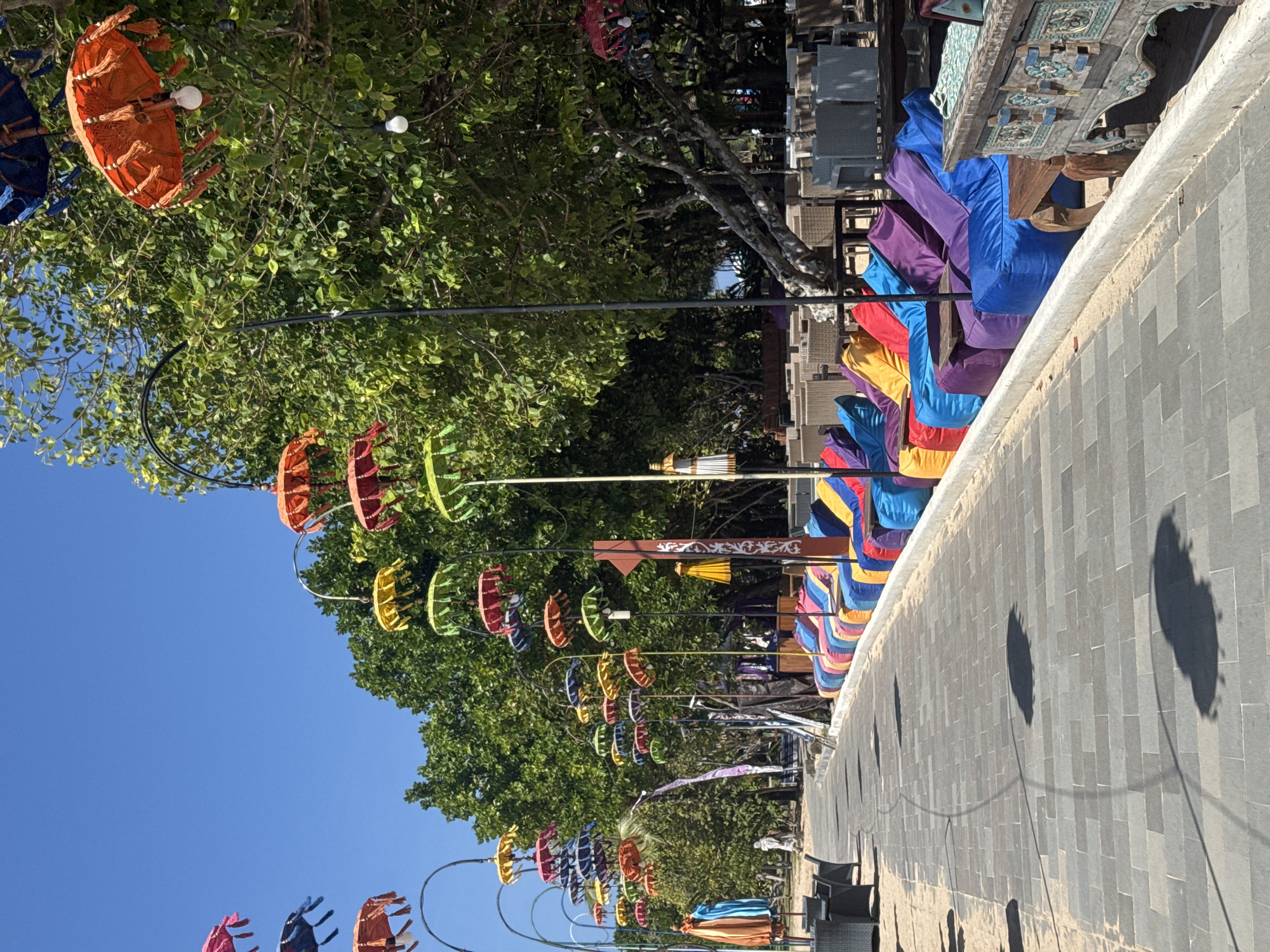 The image shows a decorated pathway lined with colorful decorations. The decorations appear to be small, umbrella-like objects in various vibrant colors such as orange, blue, purple, yellow, and green. These are strung along black wires, creating a festive and cheerful atmosphere. Some of these decorations are hanging from trees, while others are suspended along the pathway.
In the background, there are trees with lush green foliage, suggesting an outdoor setting, possibly in a tropical or warm climate. Adjacent to the pathway is a paved area with dark gray square tiles. On one side of this paved area, there are several colorful, cushioned seats or beanbag chairs, predominantly in shades of blue, purple, and red, neatly arranged. Further back, there are more outdoor furniture pieces, resembling tables and chairs made of woven material, and a decorative wooden cabinet with intricate carvings.
The sky is a clear, bright blue, indicating a sunny day. The shadows cast on the tiled pathway suggest it is midday or early afternoon. There are no people visible in the image, but the setup implies a place designed for relaxation or gathering, such as a resort, a market, or an outdoor cafe. The overall mood is bright, inviting, and festive. No visible text is present in the image.