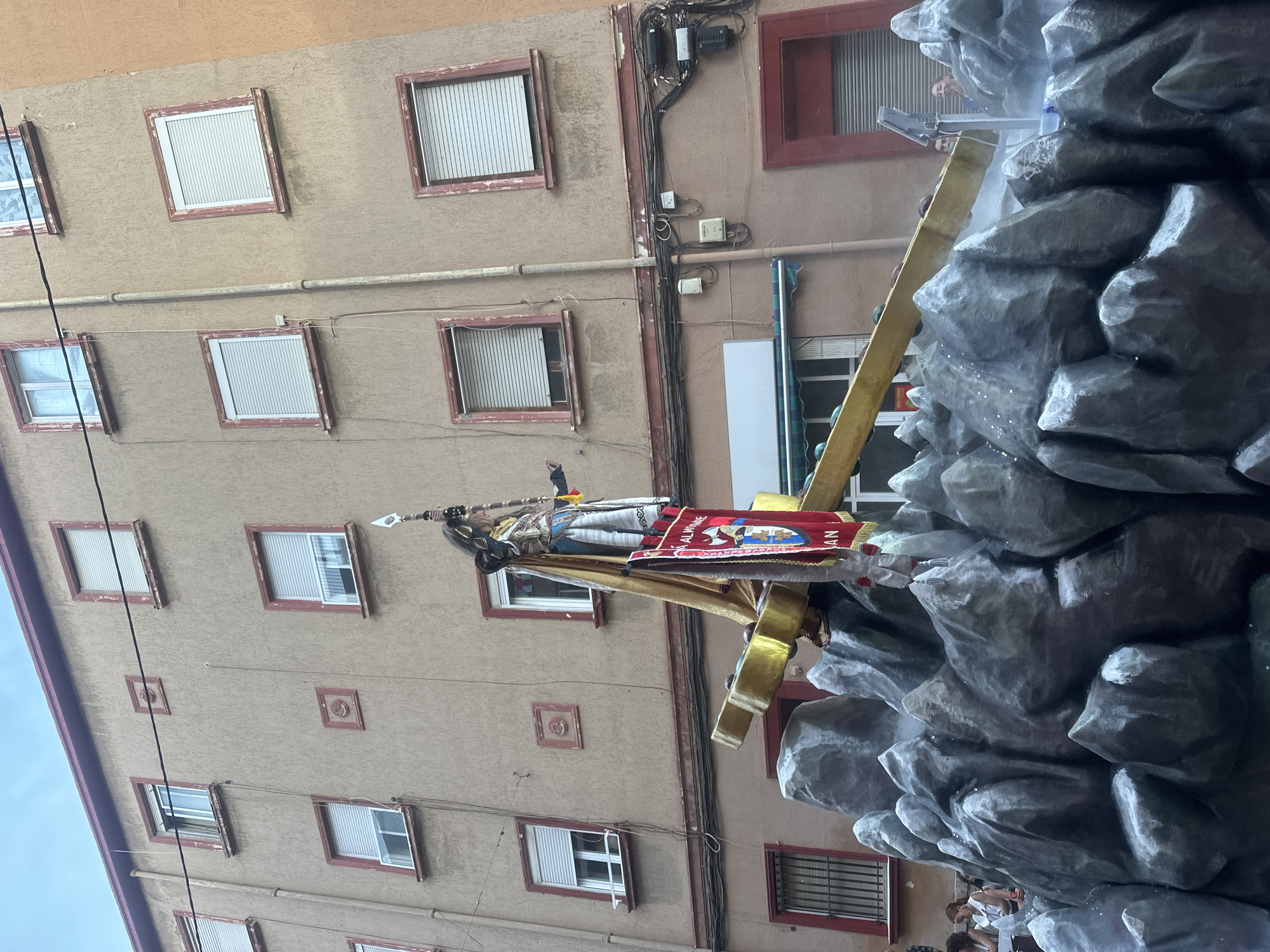 The image shows a religious float with a figure dressed in armor, holding a spear, and wearing a golden cape. The float is decorated with artificial rocks and a golden cross. A red banner with an emblem and text is hanging from the float. The float is in a street in Alicante, Spain, with buildings visible in the background. People are gathered on the street, watching the procession. The sky is overcast. The scene appears to be part of a religious festival or parade.