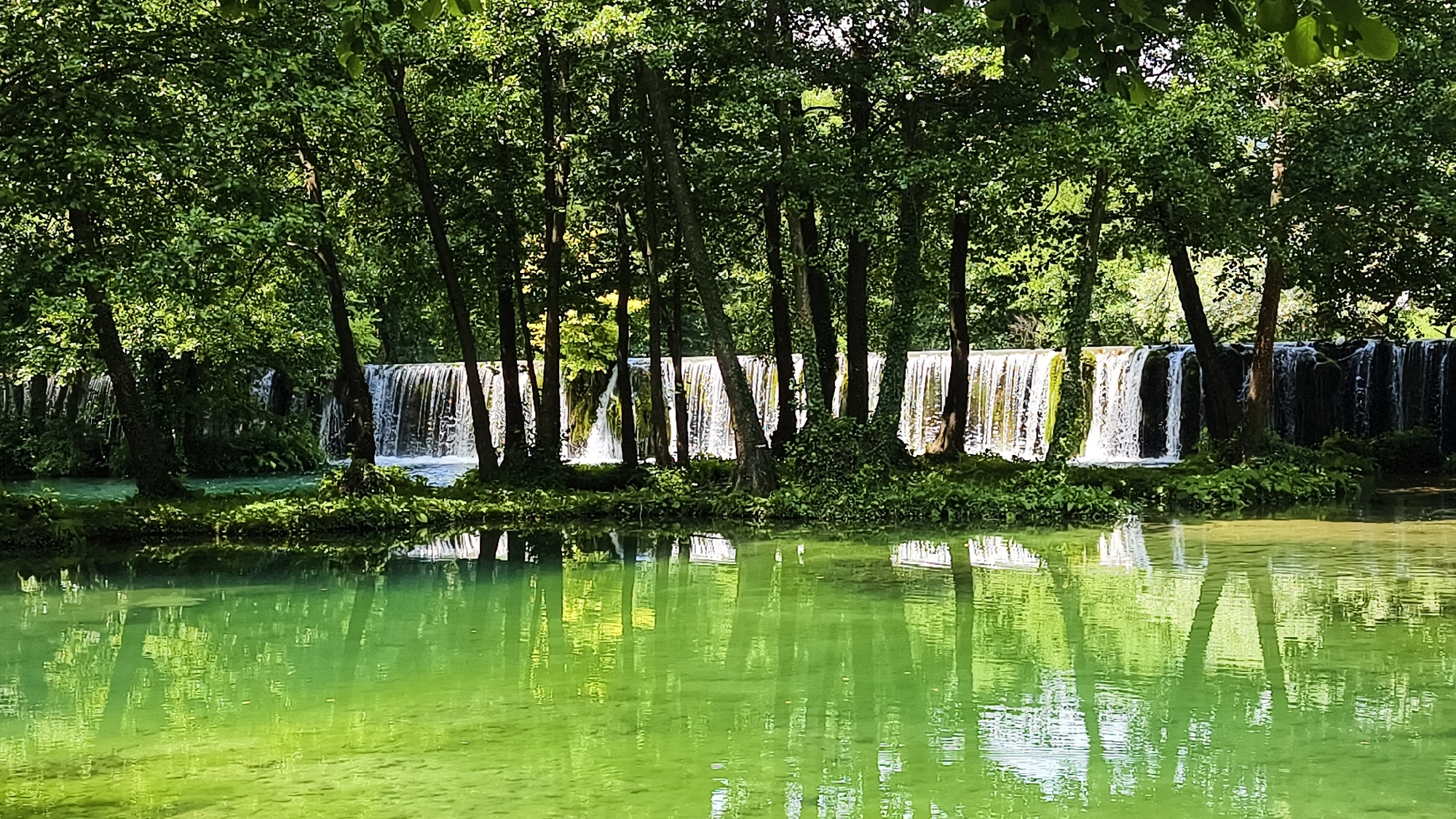 The image shows a tranquil natural scene featuring a waterfall in Vrbica, Bosnia and Herzegovina. A lush green forest surrounds the waterfall, with trees lining the banks of a clear, reflective body of water in the foreground. The water is a vibrant emerald green and mirrors the trees and the cascade, creating a serene and picturesque landscape. The waterfall itself is a wide, low cascade with water flowing over a rocky ledge, creating a curtain of white spray. The overall impression is one of natural beauty and peacefulness.