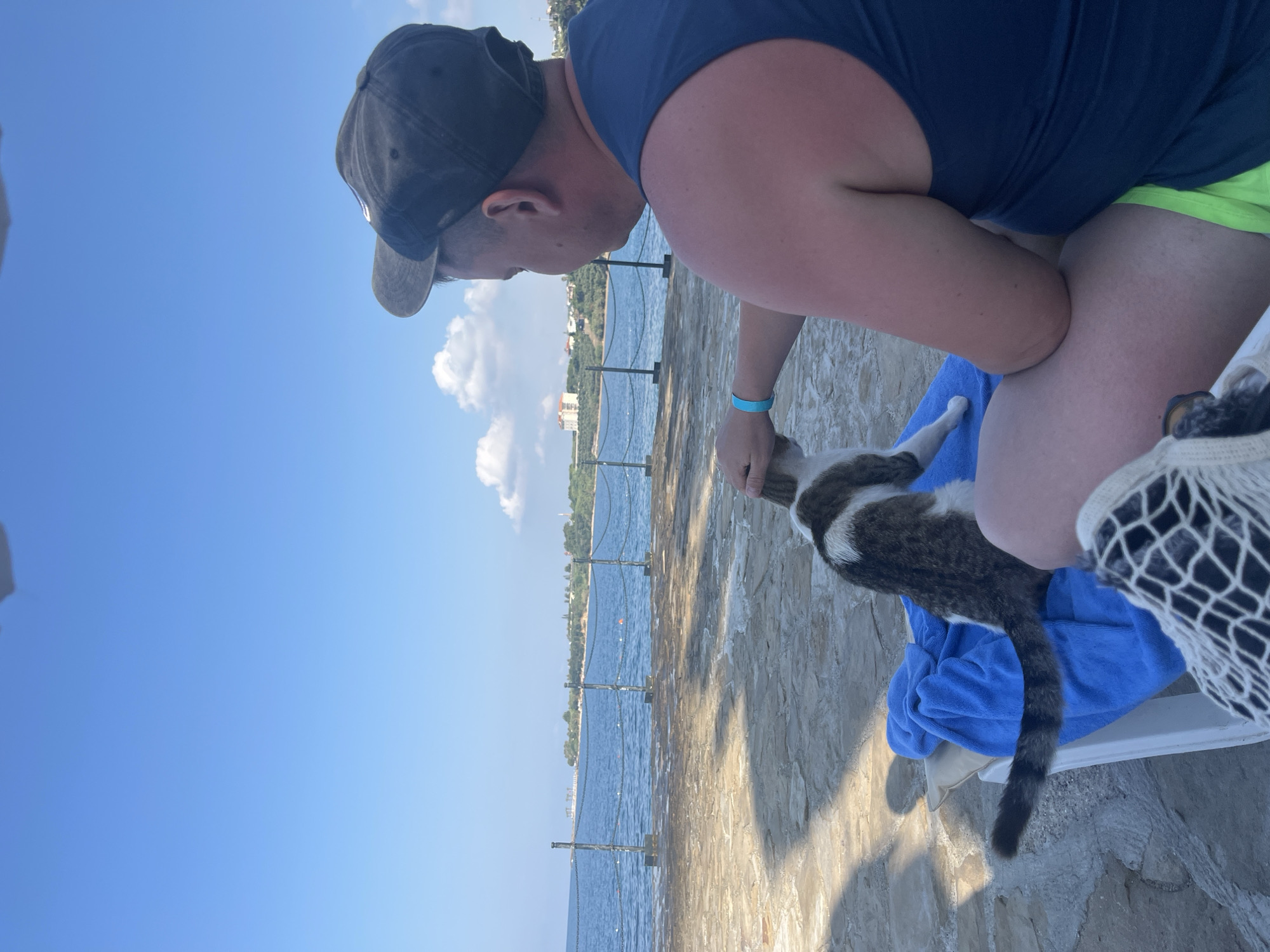 A man wearing a blue tank top and a baseball cap sits on a white lounge chair on a paved stone area near the sea. He is petting a tabby and white cat that is sitting on a blue towel. The cat is facing away from the camera. In the background, the sea is visible with a fence line separating it from the paved area. Buildings and greenery can be seen on the coastline in the distance, under a clear blue sky with scattered clouds. The location is identified as Alanya, Turkey.