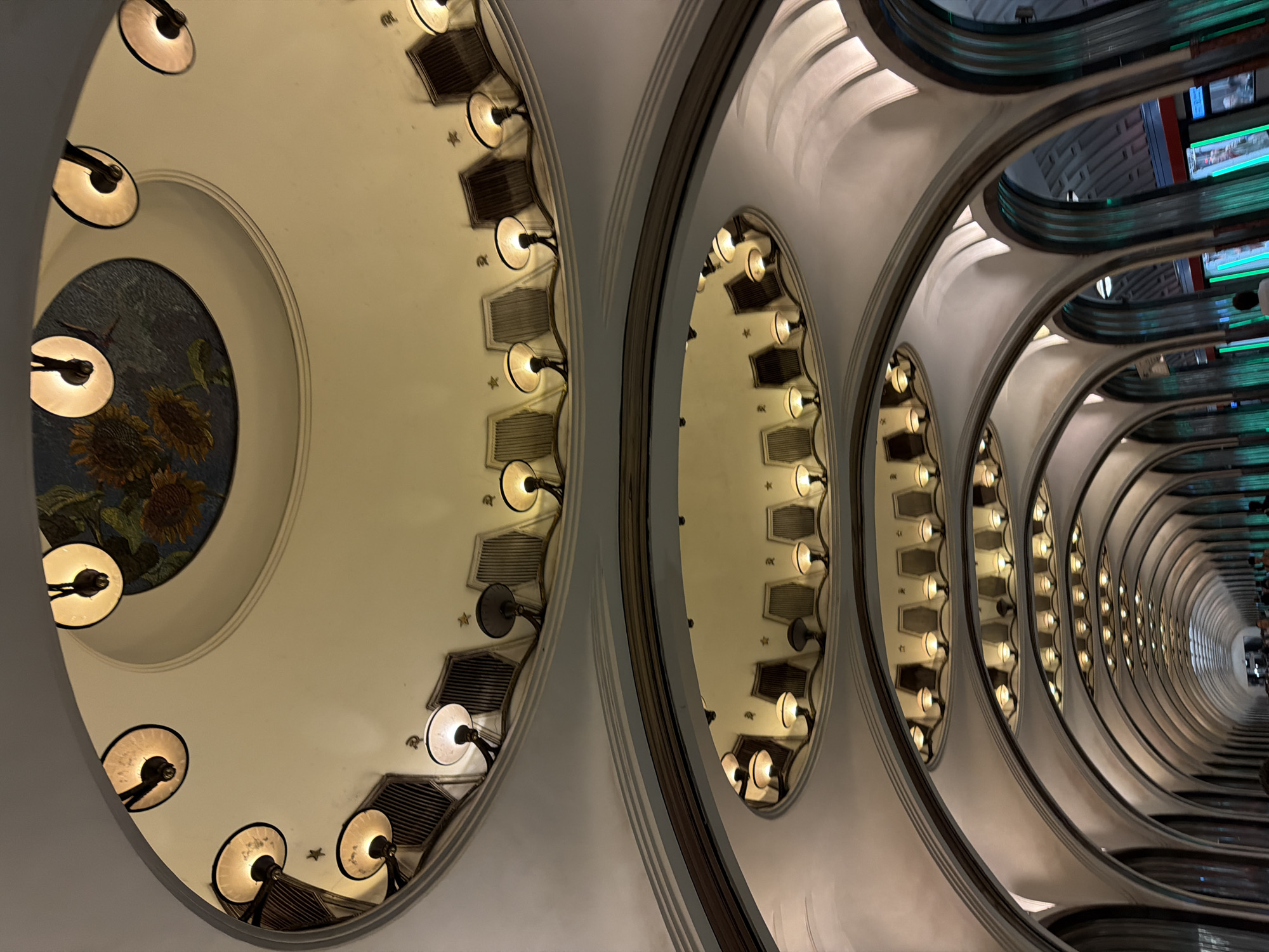 The image presents a striking upward view inside a grand, ornate escalator tunnel, characteristic of a deep-level metro or subway station. The perspective emphasizes the impressive length and architectural complexity of the structure.
On the left side, a large, circular mosaic depicting sunflowers in warm hues of yellow and brown, set against a blue-green background, is embedded in the ceiling. Surrounding this mosaic, and continuing along the curved ceiling to the right, are numerous circular light fixtures with opaque, dish-shaped shades that emit a soft, warm glow. These lights are mounted on the wall segments between repeating arched structural elements. Interspersed among the lights are decorative, angular grilles or vents, along with small, star-shaped embellishments.
The right portion of the image shows the long tunnel of the escalators, receding into the distance. The ceiling above the escalators features a similar pattern of recurring arched sections, each illuminated by multiple light fixtures, creating a rhythmic and tunnel-like effect. The metal handrails and side panels of the escalators reflect the warm light from the ceiling, as well as distinct green and blue lights from what appear to be modern digital screens or advertisements located far down the tunnel.
The scene is devoid of prominent human subjects, with only a few indistinct figures faintly visible at the very far end of the escalator, highlighting the monumental scale of the architecture. The lighting is entirely artificial, preventing any determination of the time of day. No readable text is visible. The overall impression is one of historical elegance and intricate design, suggesting a public space of significant architectural heritage.