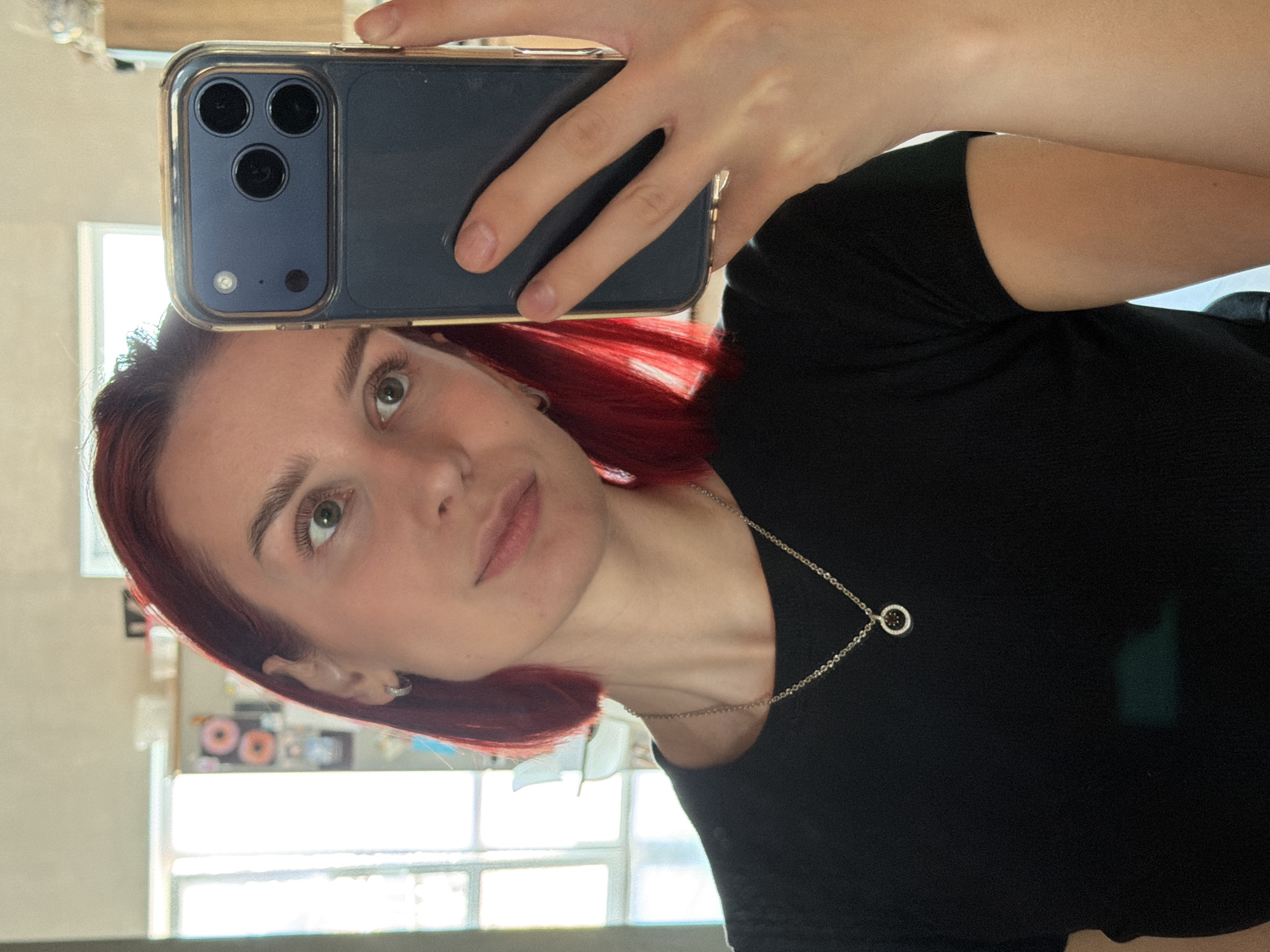 A mirror selfie, rotated 90 degrees counter-clockwise, captures a young individual looking directly into the camera. The person, likely female, has shoulder-length reddish-brown hair and light-colored eyes, appearing green or hazel. She has defined eyebrows and a neutral to slightly contemplative expression. She is wearing a black top or t-shirt and a delicate silver-colored chain necklace with a small, open circular pendant that features a dark center detail. A small silver hoop earring is visible in her left ear.
She holds a dark blue/gray smartphone, encased in a clear protective cover, horizontally at eye level. The phone is identifiable as an iPhone 12 Pro or 13 Pro series due to its prominent triple camera module, flash, and LiDAR sensor arrangement on the back. Her fingers are visible wrapped around the device.
The setting is indoors, with bright natural light illuminating the scene, suggesting daytime. A window with a horizontal pane, reflecting bright outdoor light, is visible in the background, behind the person's reflection. Other indistinct reflections, possibly of room decor or objects, can be seen in the mirror. There is no visible text in the image. The image provides no visual cues specific to Chaikovsky, Russia.