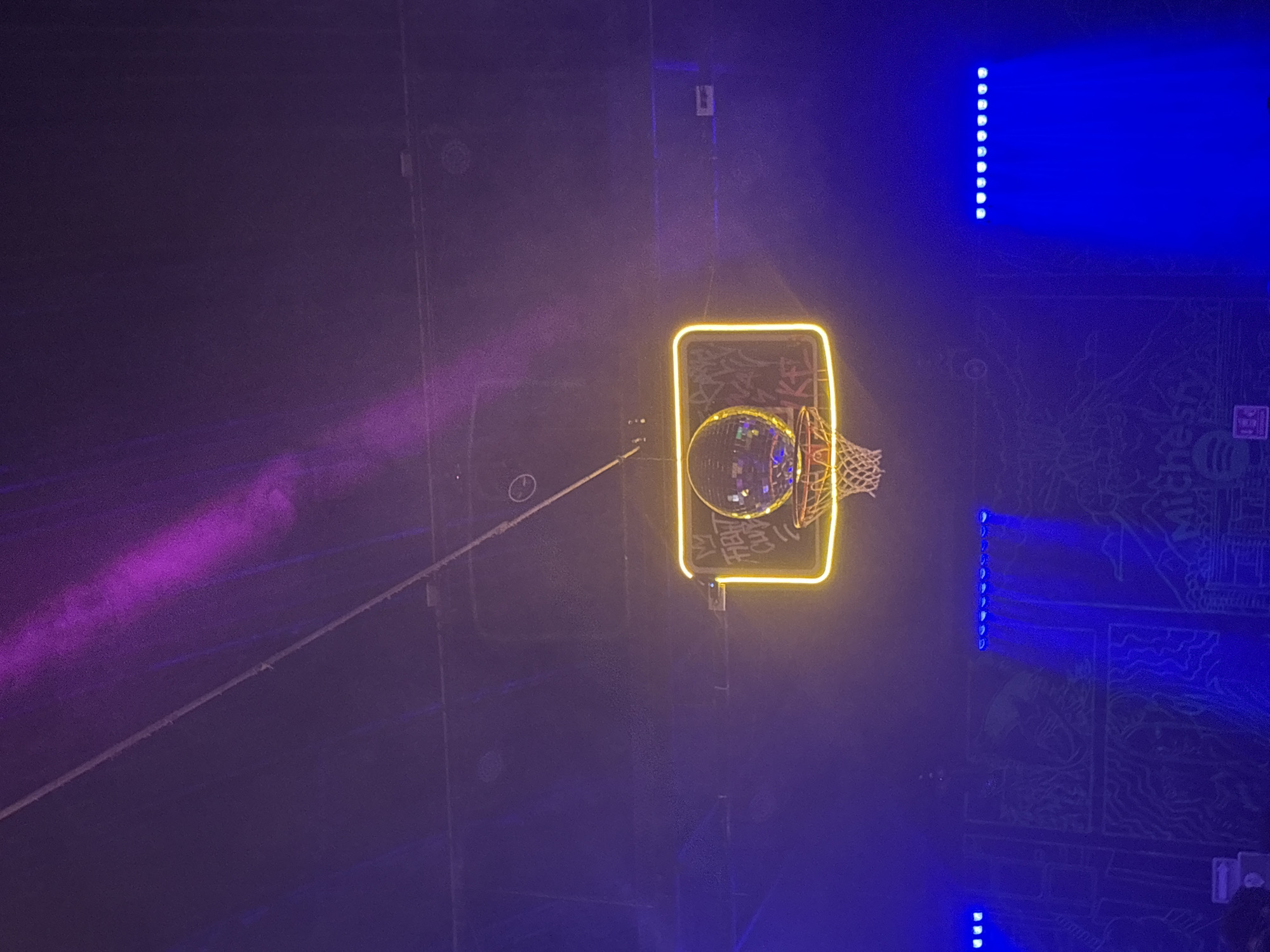 This media file presents an overhead view of a dimly lit, vibrant indoor setting, likely a bar, club, or event space in Puebla, Mexico. The main subject is a basketball hoop, creatively transformed into a decorative centerpiece. The backboard of the hoop is dramatically outlined by bright, square neon yellow lights. A sparkling disco ball is suspended from the hoop, replacing a basketball and reflecting light, while the white net hangs below.
The scene is awash in artificial light. To the left, a diagonal beam of purple light or smoke adds to the atmospheric glow. On the right, multiple vertical strips of blue LED lights brightly illuminate a section of a wall, casting a deep blue hue across it. This wall features an intricate, etched, or painted pattern. Part of the word "Michelada" is distinctly visible near the top right of this patterned wall, hinting at a Mexican-themed venue or beverage offering. Some stylized, reflective text is also faintly discernible on the basketball backboard, but it is not clearly legible.
The floor beneath the hoop appears dark and unadorned. No people are visible, but the striking combination of the disco ball, neon basketball hoop, and colorful lighting suggests a lively, festive event or nightlife atmosphere taking place during the night. The overall impression is one of a unique and modern entertainment space.