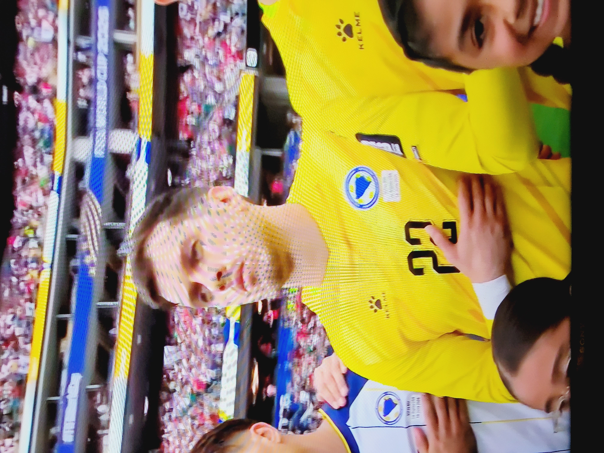 The image shows a close-up of a soccer player in a yellow jersey with the number 22 on it, standing in front of a crowd at a stadium. The player has his hand over his heart and appears to be singing or listening to a song. The crowd is blurred in the background, but it is clear that there are many spectators. A blue and yellow banner in the background reads "FIFA WORLD CUP 2026". The player's jersey has a badge on the left chest and the brand "KELME" logo on the right chest and sleeve. Another person in a white and blue jersey stands to the left of the player, and a young girl with dark hair is visible in the lower right corner, smiling at the camera. The green grass of the soccer field is visible at the bottom of the image. The location is identified as Baku City, Azerbaijan, likely at the Tofiq Bahramov Stadium.