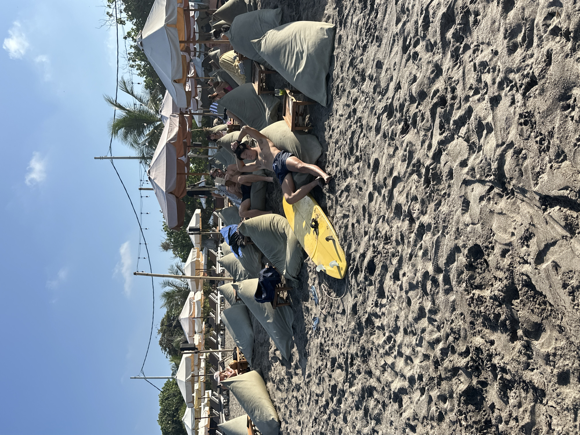 A group of people relax on a beach in North Kuta, Indonesia, with numerous large, olive-green beanbag chairs spread across the dark sand. In the foreground, a yellow surfboard rests on the sand beside a man in blue shorts and a blue shirt. A second man, shirtless and wearing blue shorts, reclines on a beanbag chair, his hands behind his head. Behind them, rows of beach umbrellas and lounge chairs are visible, with more people scattered among the beanbag chairs. Palm trees and lush green foliage border the beach, against a clear blue sky.