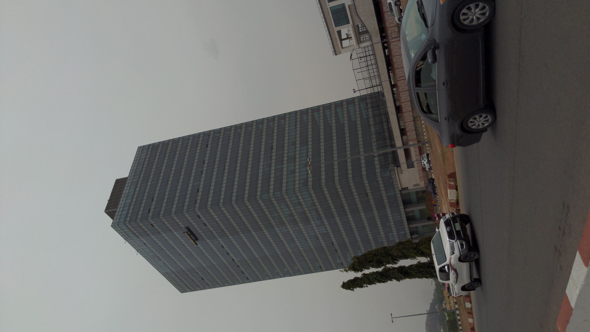 This media file captures an outdoor urban scene dominated by a tall, modern building under an overcast sky. The image is rotated, but when reoriented, it shows a multi-story, contemporary skyscraper with a patterned glass and panel facade on the left. The building's architecture features a grid of windows and a stepped or receding profile on one side.
A paved road stretches across the foreground and right side of the frame. On the road, two cars are visible: a white SUV or pickup truck closer to the viewer, and a dark-colored sedan further down the street. Concrete road barriers, marked with red and white stripes, line the edge of the road. Faint human figures can be observed near the white vehicle, suggesting people are present.
Beyond the road, some green trees and other structures are visible, and in the distant background, the subtle outlines of hills or mountains can be seen beneath the hazy, light grey sky. The lighting suggests daytime, possibly a cloudy day or a time with soft, diffused light. There is no legible text visible on any buildings, vehicles, or signs. The scene depicts a modern urban environment, consistent with a city like Abuja, Nigeria.