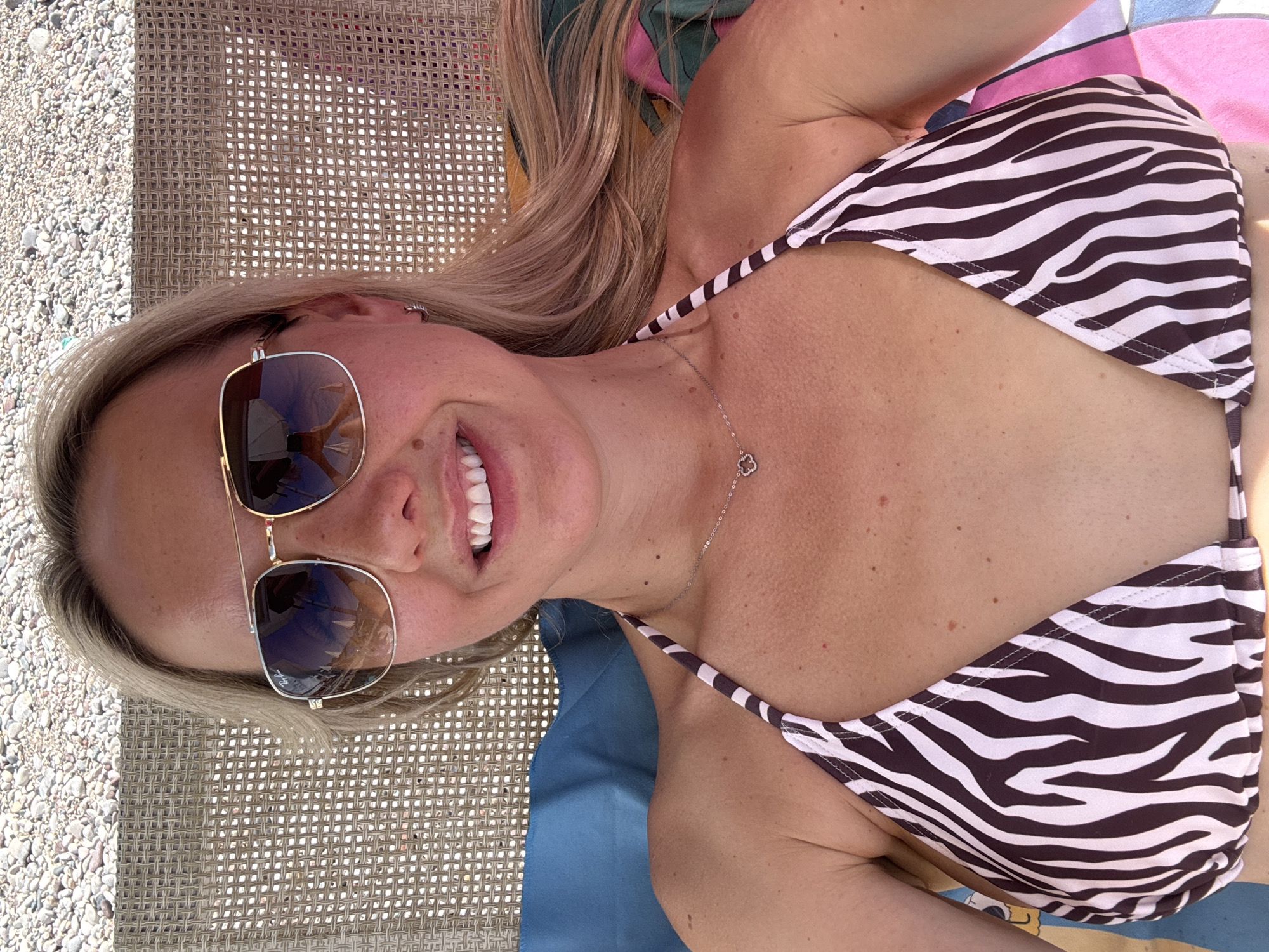 A woman is lying on a sun lounger on a pebble beach, likely in Kamenovo, Montenegro. She is wearing aviator sunglasses and a brown and white zebra-print bikini top. She has blonde hair and is smiling at the camera. The background shows a blurry expanse of pebbles and the woven material of the sun lounger.