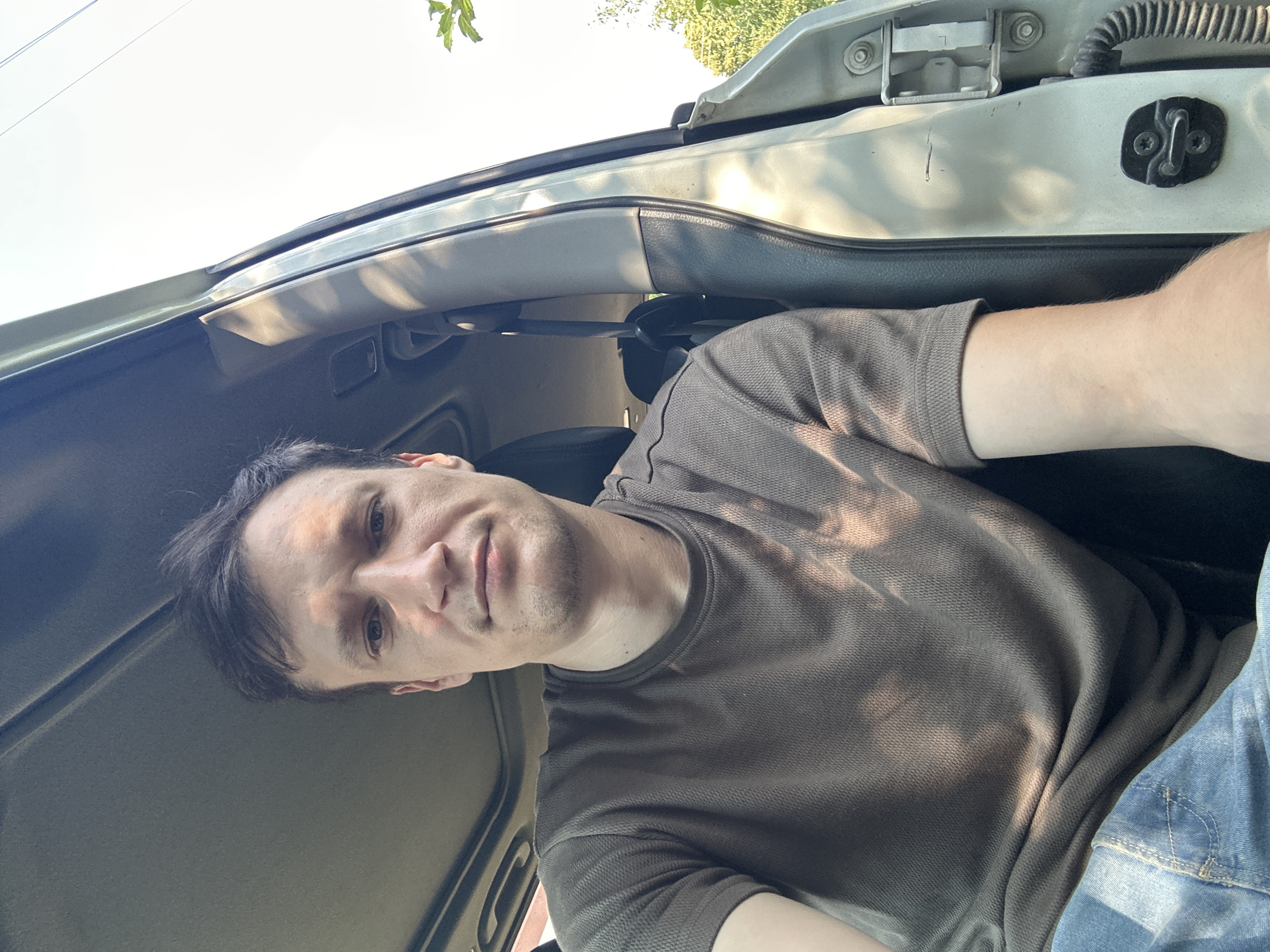A selfie of a man sitting in the driver's seat of a car with the door open. The man has short, dark brown hair, fair skin, and a light stubble. He is wearing a brown t-shirt and blue jeans. The interior of the car is visible, including the sunroof and the B-pillar. Outside the car, trees and a clear sky are visible. There are no distinguishing landmarks to place this image in Shatava, Ukraine.