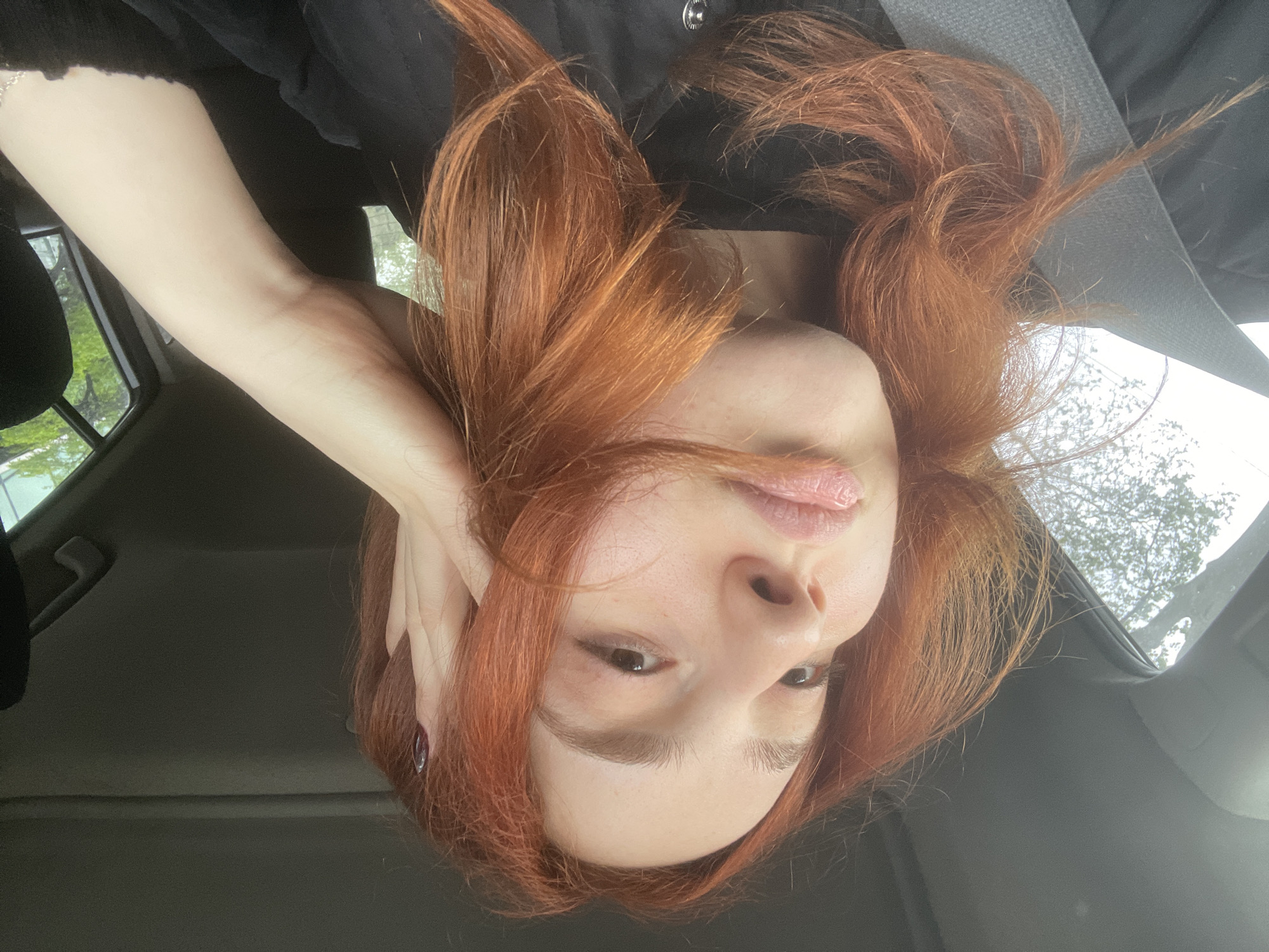 A young woman with red hair is sitting in a car. She is wearing a black top and a seatbelt. Her left hand is touching her head. The car is parked, with trees visible through the window. The interior of the car is gray. The location is likely Tbilisi, Georgia, though there is no definitive evidence to confirm this in the image.