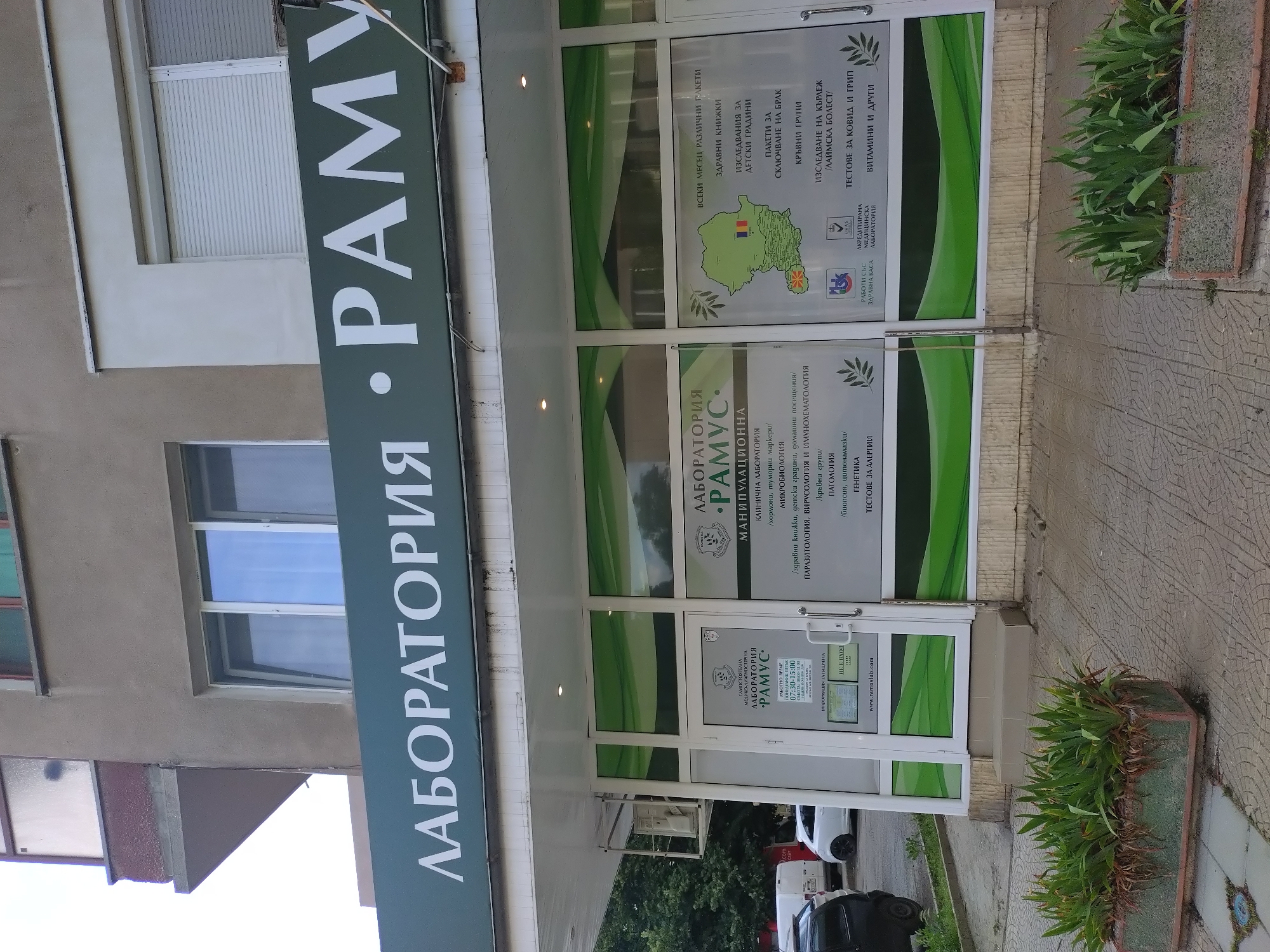 The image shows the exterior of a laboratory called "ЛАБОРАТОРИЯ РАМУС" (Laboratory Ramus). A dark green sign with white lettering displays the laboratory's name vertically on the left. The entrance consists of glass doors and windows with green and white accents. The windows display information about services offered, including clinical laboratory services, microbiology, parasitology, genetics, and tests for COVID and flu. A sign on the door indicates operating hours and contact information. Potted plants flank the entrance. The location is assumed to be in Stara Zagora, Bulgaria, based on the Cyrillic script used in the signage. Several cars parked along the road can be partially seen in the background.