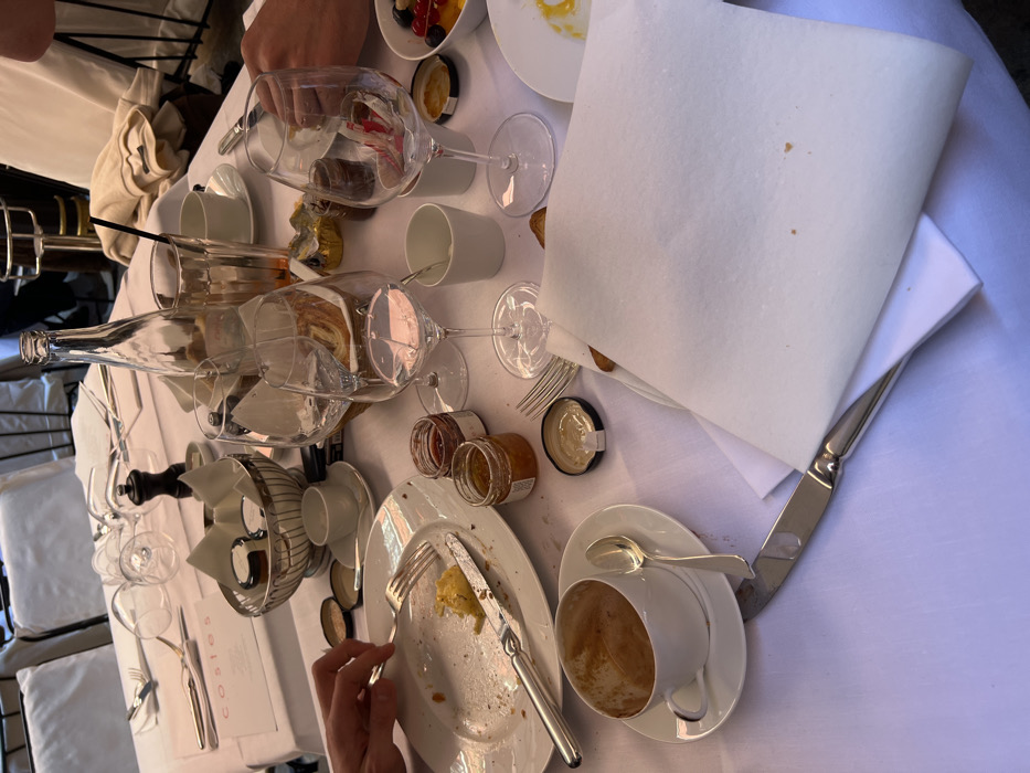 The image displays a dining table after a breakfast or brunch meal at Hotel Costes, located at 239 Rue Saint Honoré, Paris, France. The table is covered with a white tablecloth.
Essential details include a white plate on the left with minimal yellow food residue, a fork, and a knife. An empty white coffee cup, saucer, and spoon are positioned below. Two small glass jars of jam, one red and one amber, are present, with an open lid near a fork. A silver bowl holds additional individual servings. A black salt and pepper grinder stands nearby.
Centrally, an empty clear glass bottle, multiple stemmed wine glasses (some containing liquid), and a tall glass with an orange beverage and straw are arranged. A white folded napkin covers a large knife in the lower right. A hand is partially visible on the far right, adjacent to a white bowl of mixed fruit and another white plate with yellow food, possibly a fried egg. A menu card on the table clearly reads "costes." The background shows blurred white-backed chairs.