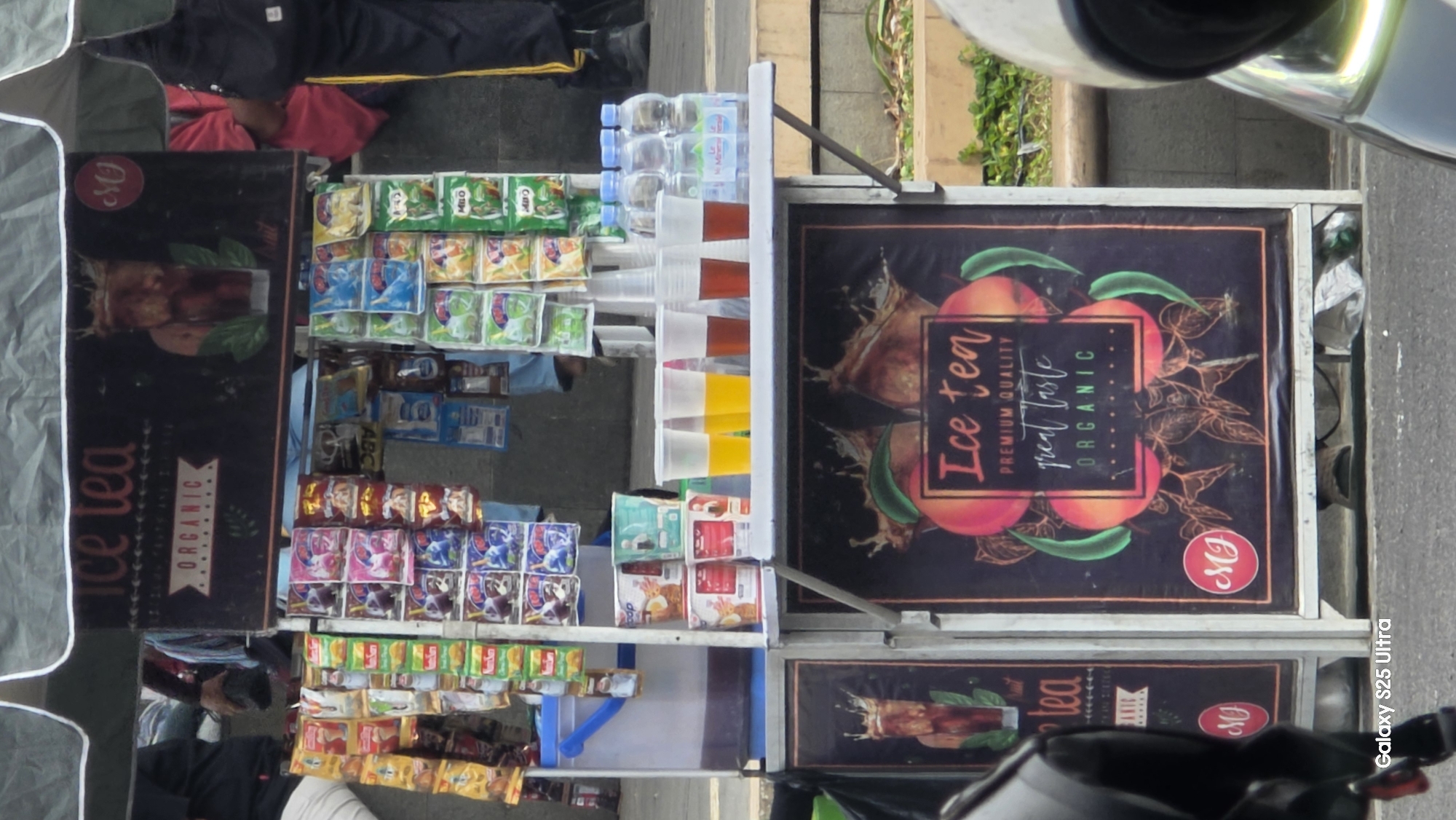 The image shows an outdoor beverage stand in an urban setting, possibly Kecamatan Pati, Indonesia. The stand is displaying various products, including packaged snacks and drinks. On the upper portion of the stand, a banner reads "Ice Tea ORGANIC" with an image of iced tea. Below this, shelves are stocked with colorful snack packets. In the middle section, there are cups filled with yellow and brown liquids, and several bottles of mineral water. The lower section features another banner with "Ice tea PREMIUM QUALITY great taste ORGANIC" and images of fruit. The stand appears to be a small business operating on a sidewalk, with people in the background, suggesting a public area. The overall scene is that of a typical street vendor setup.