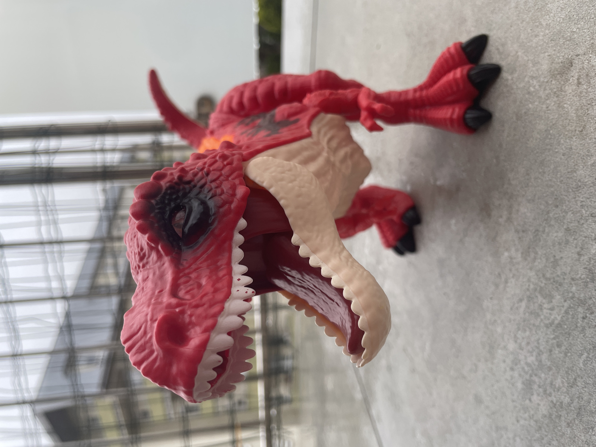 A close-up, eye-level, outdoor shot shows a bright red toy dinosaur, likely a Tyrannosaurus Rex, on a gray tiled surface. The dinosaur's mouth is wide open, revealing a pink tongue and rows of white teeth. Its eyes are black with some darker markings around them. The dinosaur has black markings on its back and orange accents near its neck. The background is blurred, showing vertical bars, likely a fence or railing, and a building structure with windows. The overall lighting suggests an overcast day. The location is unknown.