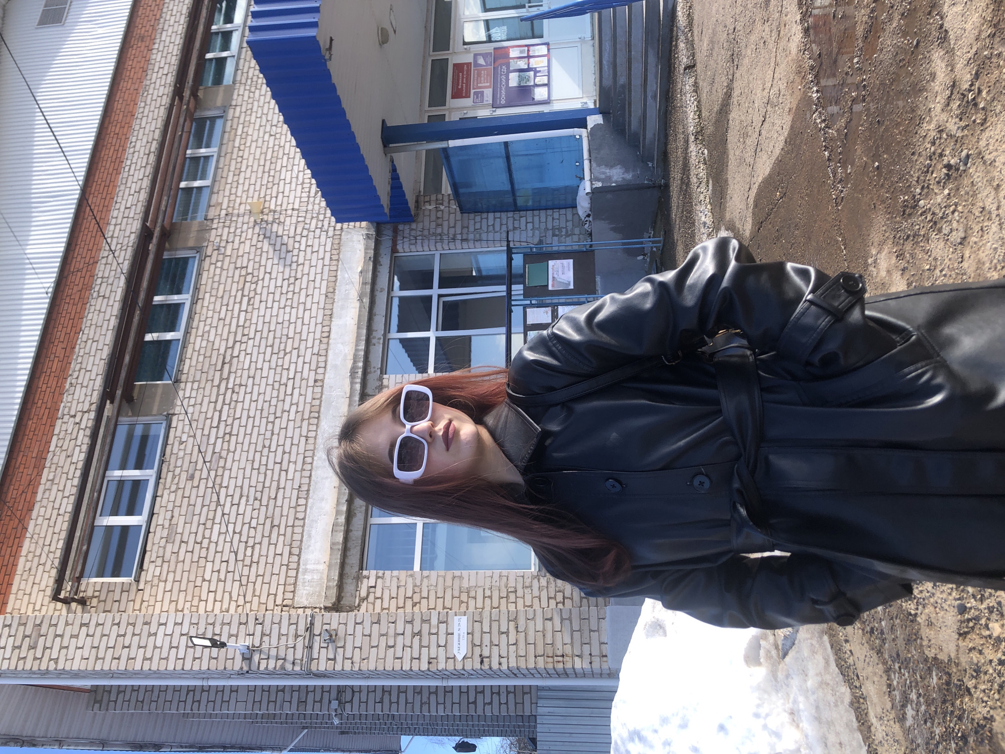 The image depicts a young female subject standing outdoors in front of a light-colored brick building in Фоки, Russia, specifically the Fokinsky House of Culture (ФОКИНСКИЙ СДК). The subject has long reddish-brown hair and is wearing a black, belt-tied, leather-look trench coat with her hands in the pockets, and distinctive white, rectangular-framed sunglasses. She is looking towards her right (viewer's left).
The building behind her is two stories high, constructed of light-colored bricks, with multiple white-framed windows. A prominent blue corrugated metal awning extends over a recessed entrance area on the right, supported by a blue pillar. Steps lead up to the entrance, which features glass doors displaying various information posters. One poster clearly reads "ФОКИНСКИЙ СДК." To the left of the entrance, an information board with several notices is visible.
On the brick wall to the subject's left, a white directional sign with a black arrow points left, displaying "УБЕЖИЩЕ № 29,25" and "130 м," indicating a shelter. A modern, dark-colored outdoor light fixture is mounted on the building wall near the corner. The ground in the foreground is uneven, composed of mud and gravel, with visible patches of melting snow and puddles. The sky is clear and blue, indicating a bright, sunny day, with shadows cast on the ground. A corrugated metal fence is partially visible on the far left.