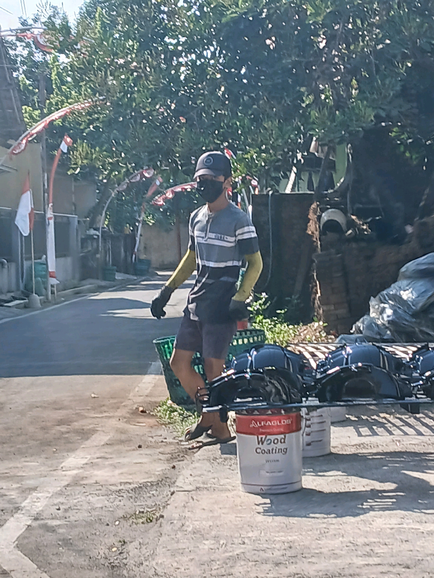 The image depicts a street scene in Jepangpakis, Indonesia. A person wearing a dark cap, a mask, and a grey and white striped shirt is walking on a paved street. They are also wearing dark shorts, yellow sleeves, and black gloves. In the background, there are trees, buildings, and Indonesian flags decorating the street. In the foreground, a metal rack holds several dark, round objects, possibly motorcycle wheels or similar items. Two white paint buckets are visible, one with a red and white label that reads "ALFAGLOS Wood Coating". The ground is paved and appears to be a road or walkway, with some patches of grass and debris. The scene is lit by natural sunlight, casting shadows on the ground.
