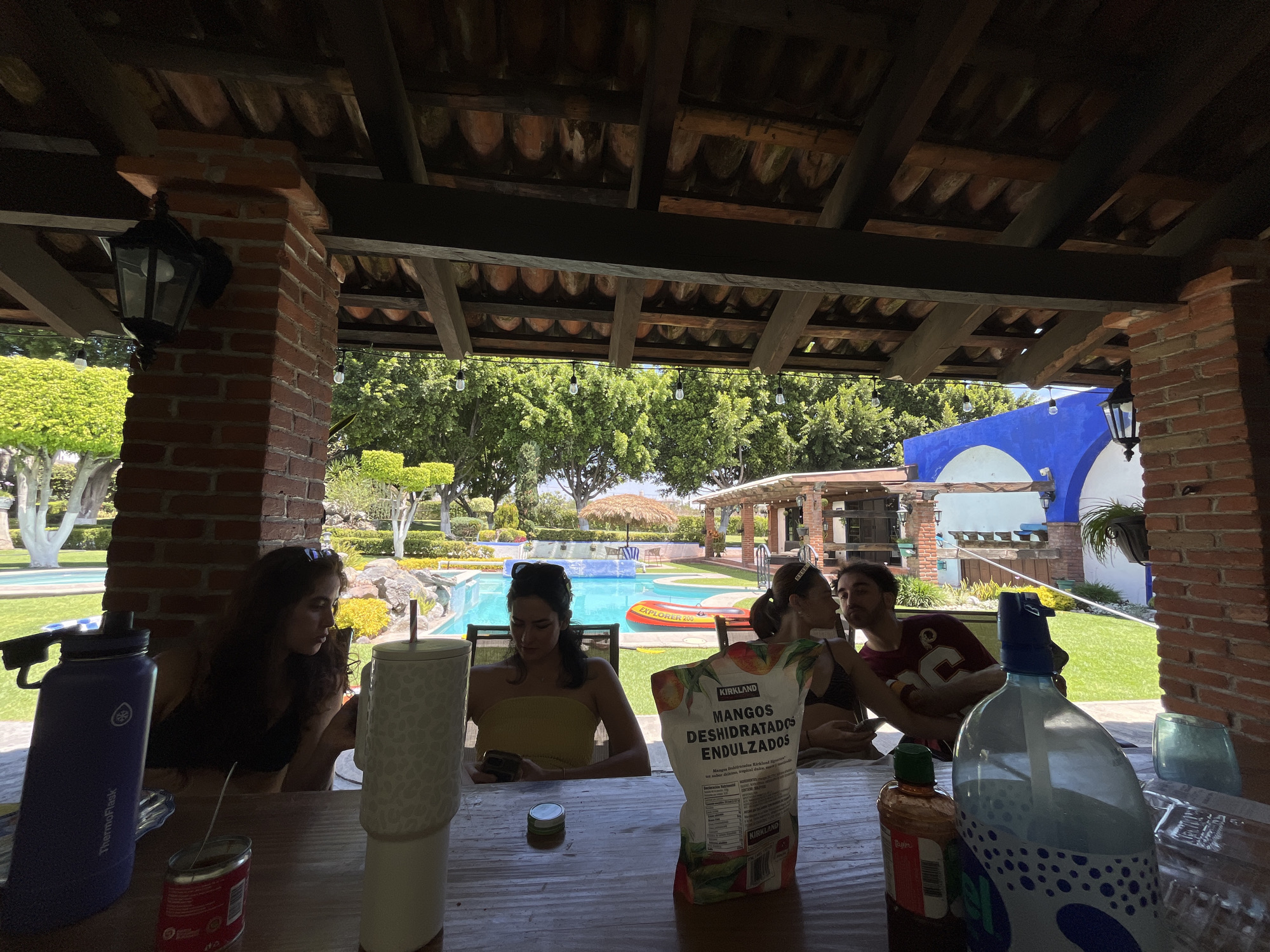 Captured from beneath a roofed structure, the image presents a scene where four individuals are seated around a table on a sunny day. The environment is characterized by a well-maintained lawn, a swimming pool, and various architectural elements.
To the left, a woman with dark hair is seated, partially obscured by a blue water bottle. To her right, a woman wearing a yellow top is looking at her phone. The table is cluttered with items including a large white cup, a bag of dried mangos, and a small jar of hot sauce. A couple sits on the other side of the table and they are looking at each other.
In the background, beyond the table, a swimming pool is bordered by lounge chairs and greenery. A tiki hut is visible, and a blue-and-white building stands further off in the distance.
The architectural elements of the covered structure, including brick columns and a tiled roof, are clearly visible. Overhead, exposed beams and tiles make up the ceiling. The setting appears to be a residential or resort-style outdoor space.