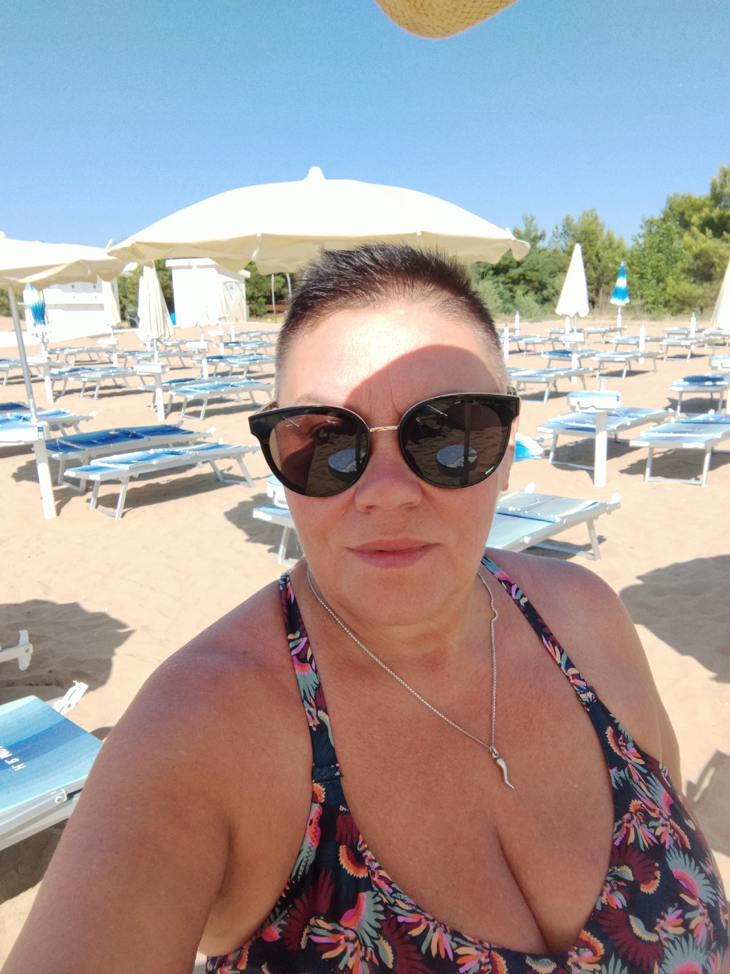 A woman is taking a selfie on a sandy beach in Peschici, Italy. She is wearing sunglasses and a floral bikini top, and a silver necklace with a chili pepper charm. The beach is lined with rows of empty lounge chairs and umbrellas, suggesting it is a public beach with amenities. The sky is clear and blue, and the sun is casting shadows, indicating it is daytime. The background shows more beach chairs, umbrellas, and some trees and vegetation.