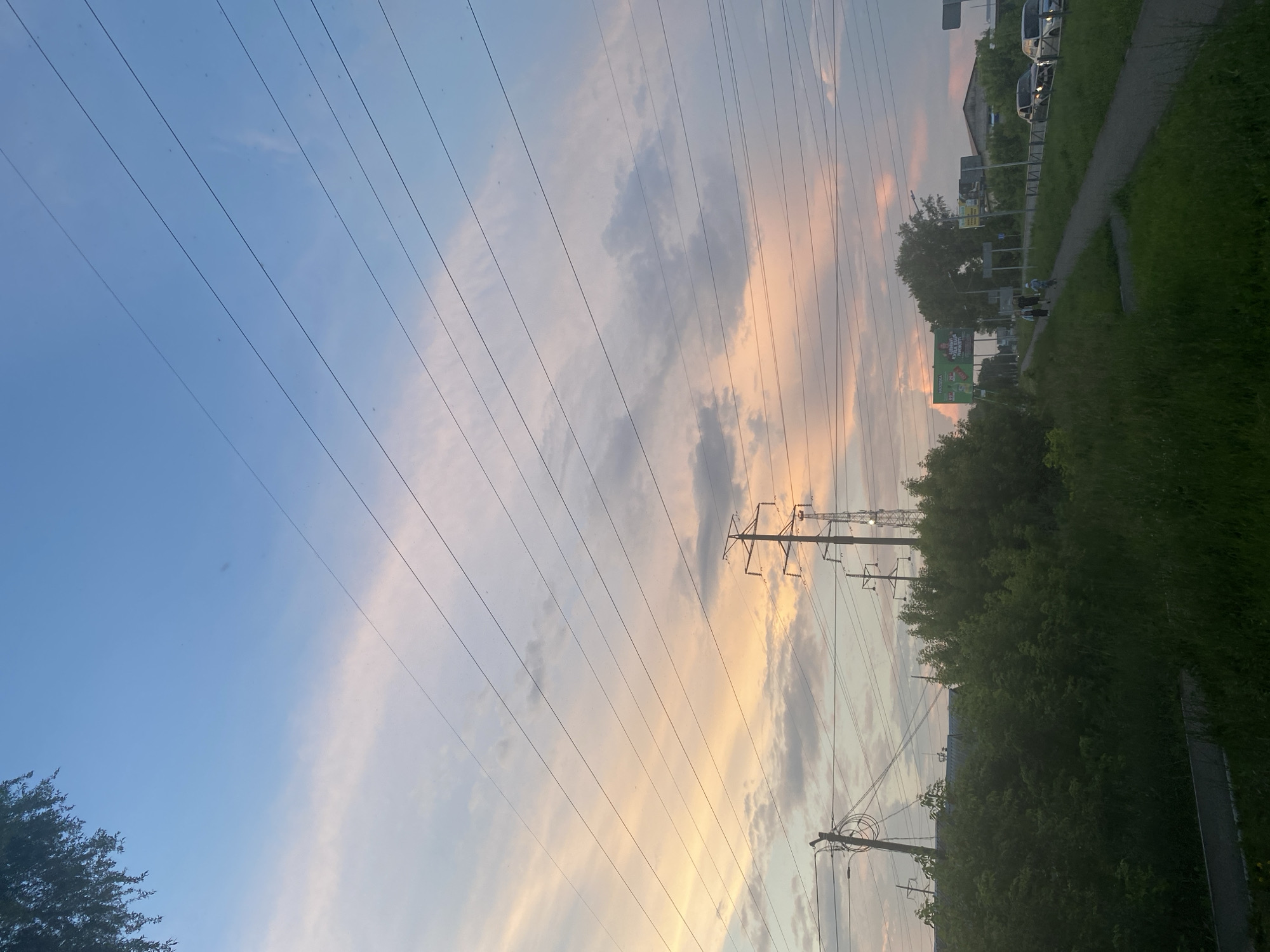 The image depicts a cloudy sky with streaks of pink and orange, suggesting either sunrise or sunset. Several power lines crisscross the frame, some supported by tall transmission towers. In the foreground, a green, overgrown area with grass and bushes lines a paved walkway. Beyond the walkway, a road is visible with a few cars and some buildings in the distance. A billboard with Russian text is also present. Two figures, a child on a bicycle and two adults walking, are on the walkway. The overall setting appears to be an urban or suburban environment.