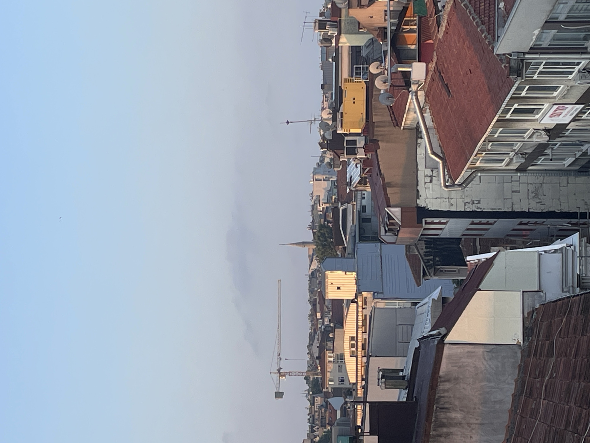 This is a medium-shot, eye-level photograph of a cityscape, likely in Kadıköy, Turkey, as indicated by the presence of a construction crane and a spire that could be part of a mosque or church. The image is dominated by rooftops of various buildings, mostly with red-tiled roofs, suggesting a densely populated urban area. Some buildings appear to be residential, while others might be commercial, given the presence of what looks like a large generator or industrial equipment on one rooftop, accompanied by satellite dishes. The sky is a pale blue with some wispy clouds, and the light suggests it is either early morning or late afternoon, casting a warm glow on some of the building facades. There are no people visible in the frame. The overall impression is one of an active city with ongoing construction and a mix of older and newer structures.