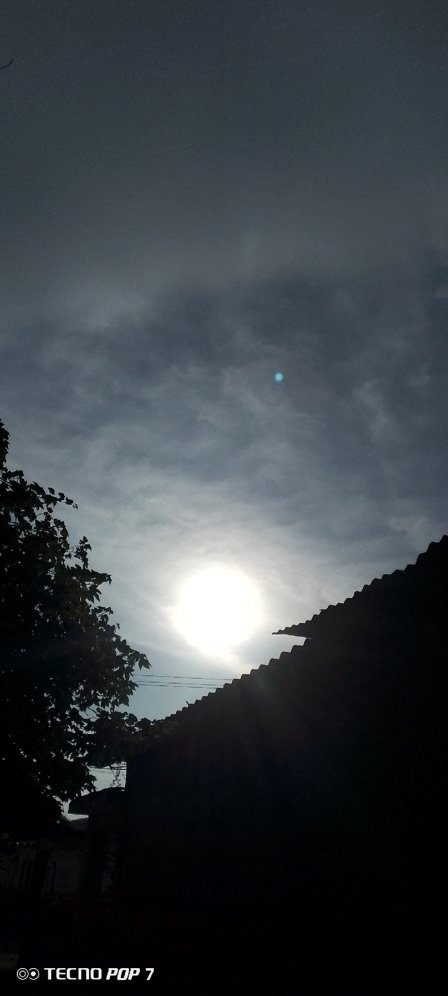 The image, taken in Altagracia, Venezuela, depicts a silhouetted rooftop against a cloudy sky. The sun is visible, shining brightly and partially obscured by the clouds, creating a halo effect. A tree with dark leaves is visible in the lower left corner, also silhouetted. The texture of the rooftop suggests it is made of corrugated material. The lower portion of the image contains a watermark indicating it was taken with a "TECNO POP 7". No people or specific actions are evident in the scene.
