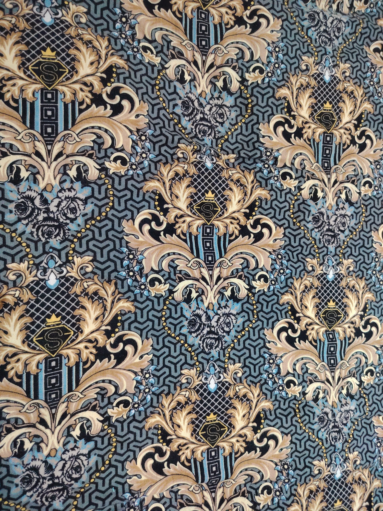 This image depicts a close-up view of a fabric with an ornate, damask-inspired pattern. The pattern features stylized floral elements in shades of gold and beige, intertwined with darker black and grey motifs. A repeating geometric background in muted teal and grey hues forms the base of the design. Within the floral arrangements, there are black diamond shapes containing a gold "S" logo topped with a small crown. Small clusters of blue and grey flowers are also present throughout the pattern. The lighting appears to be even, suggesting an indoor setting or overcast conditions. There is no indication of the city of Pitnak, Uzbekistan, or any specific environmental context within this image.