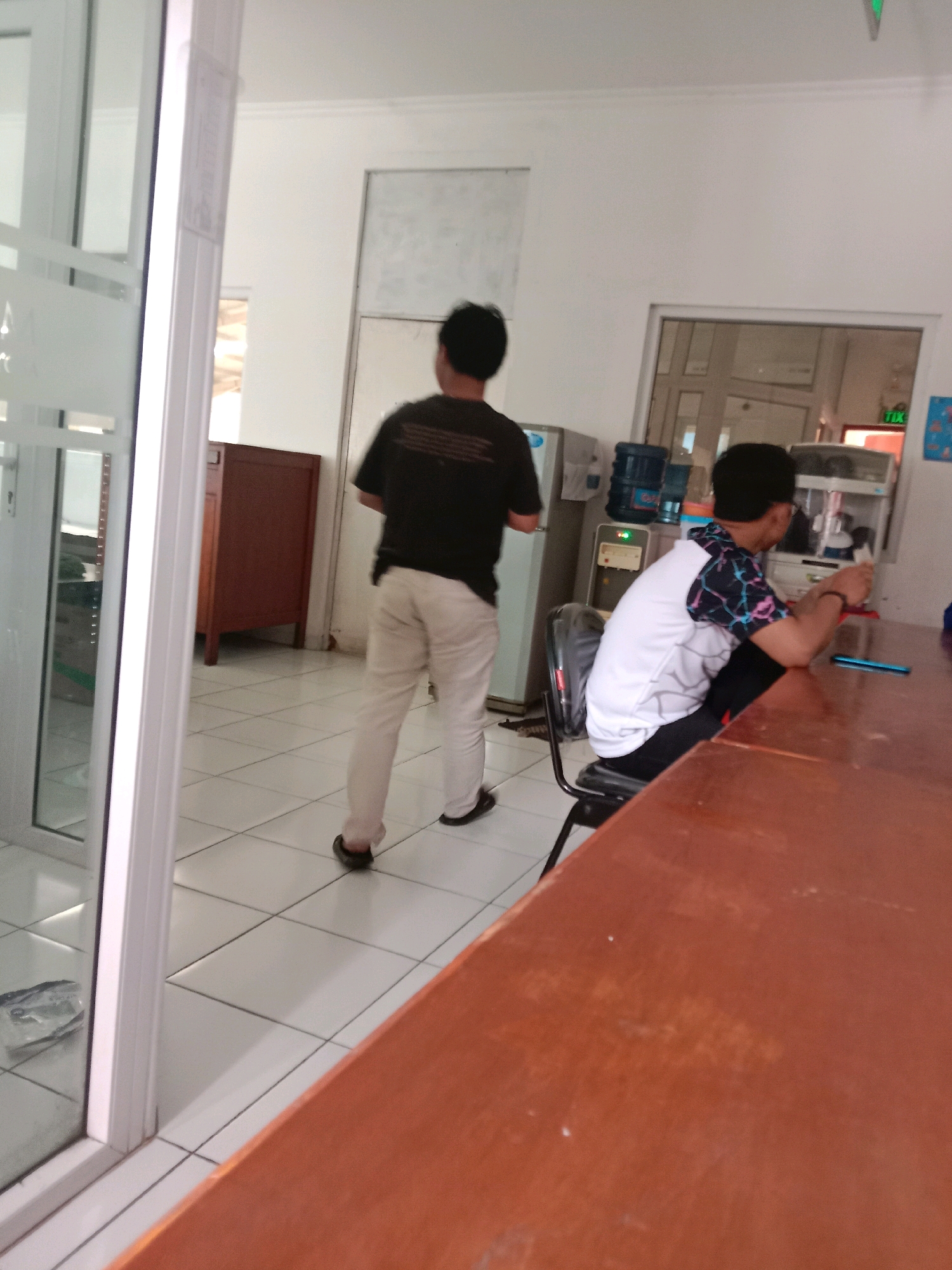 The image depicts an indoor scene, likely an office or communal area, with elements suggesting a location in Semarang, Indonesia. In the foreground, a light brown, polished wooden table dominates the lower right quadrant. To the left, a person wearing a white t-shirt with a black, blue, and pink geometric pattern on the sleeves and shoulders sits on a black office chair, facing away from the camera and towards the table. They are wearing dark pants and have their right hand resting on the table, possibly holding a light blue object, which could be a phone.
Further into the scene, a person in a black t-shirt and light-colored pants walks away from the camera towards the left. Their posture suggests they are moving from one area to another. Behind them, a wooden cabinet with two drawers is visible. In the background, there are white walls, a water dispenser with two blue water bottles, and a glimpse of what appears to be a whiteboard or a large printed notice on the wall. The floor is tiled with white square tiles. A glass-paneled door or window is on the far left, revealing a reflection of the interior. The overall lighting suggests an indoor environment, possibly during the daytime. The presence of office furniture, a water dispenser, and the general layout are consistent with common workplace settings in Semarang.