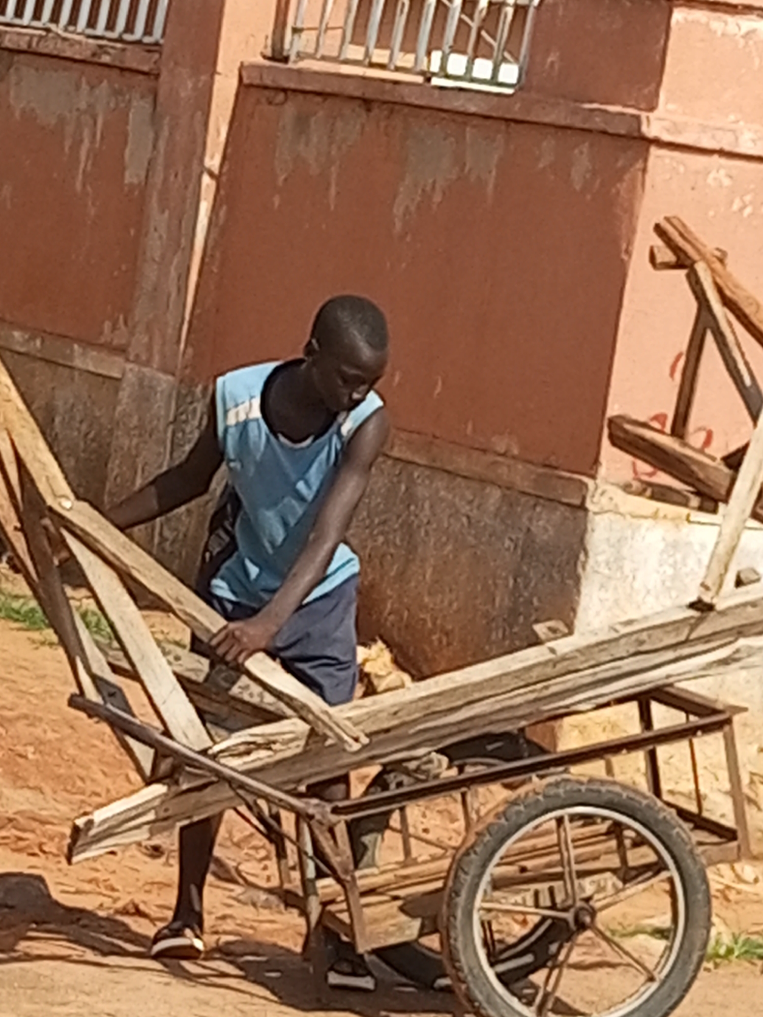 A young Black male is standing by a wooden cart with a metal frame and one wheel. He is wearing a light blue sleeveless shirt with white trim and navy blue shorts. His right arm is extended, resting on a piece of wood on the cart, and he appears to be holding another piece of wood. The cart is filled with several long pieces of wood. The ground is dirt and sand. Behind the cart is a building with a salmon-colored wall and a white barred window. The scene is outdoors in daylight.