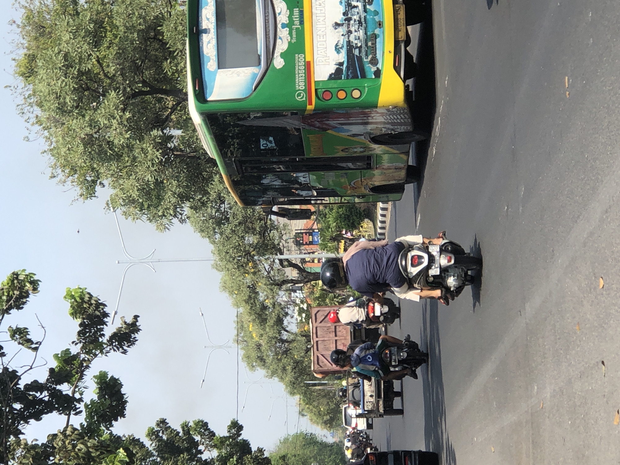 A sunny daytime, outdoor, eye-level, medium shot captures traffic on a paved road in Larangan, Indonesia. A bright green and yellow bus, emblazoned with "Trans Jatim" and images of boats and a cityscape, is prominently positioned on the right side of the frame. The bus has several passengers visible through its windows and displays a phone number, "0811356500," and the text "LAYANAN PENGADUAN." Further left on the road, a small truck with a brown cargo bed is visible, followed by several motorcyclists. One motorcyclist in the foreground, wearing a blue shirt and helmet, is riding a scooter. Another motorcyclist ahead of him wears a red helmet. The road is lined with trees and streetlights, and the sky is clear and blue. The scene depicts typical urban road activity.