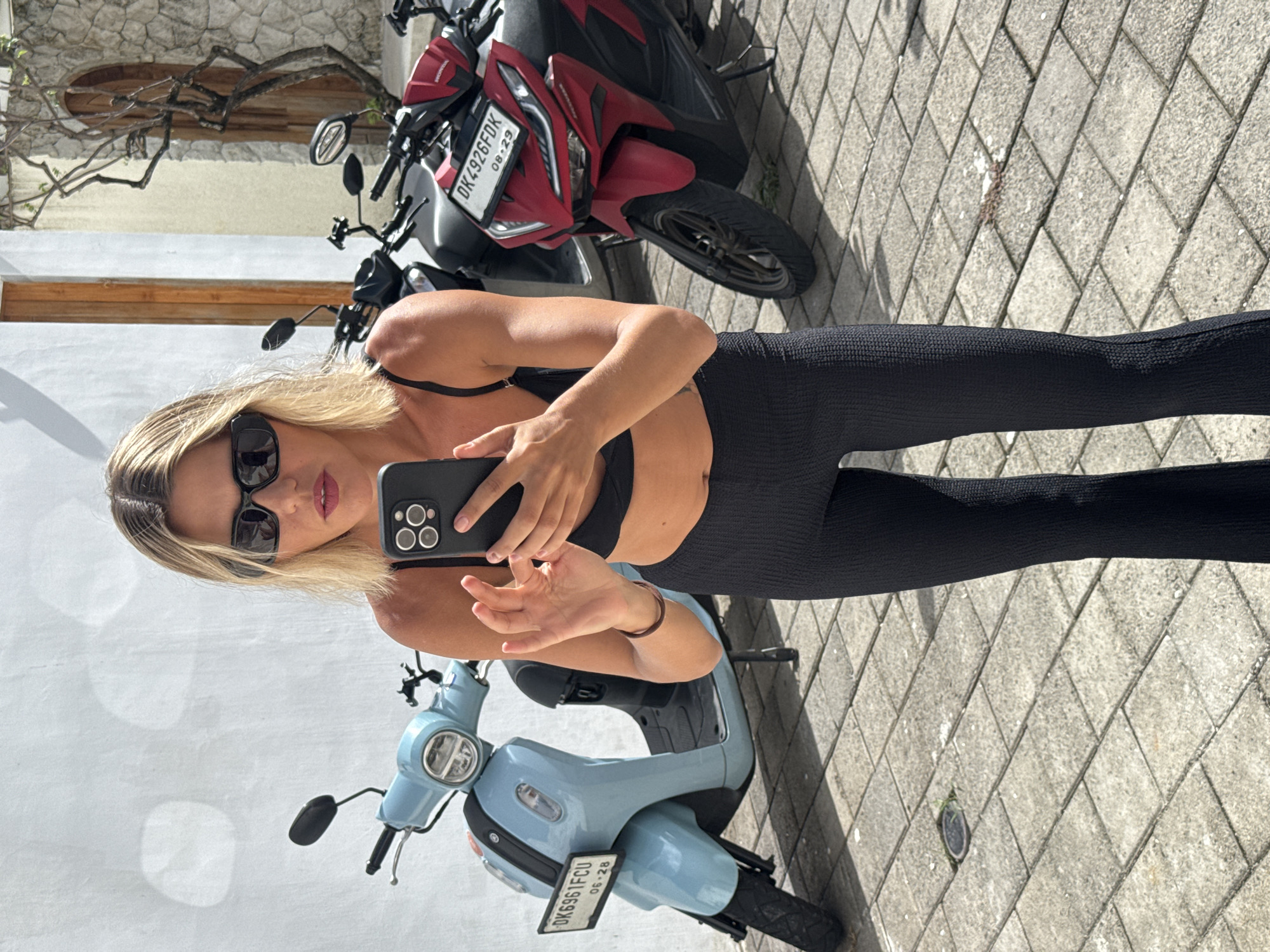 The image depicts a woman standing on a stone-paved surface alongside two parked scooters. She is wearing a black bra top and leggings, sunglasses, and holding a black smartphone in her hand, facing the camera. The scooter closest to her is light blue, with a license plate reading "DK6961FCU." Behind it is a red motorcycle with a license plate reading "DK4926FDK." A light-colored building with visible architectural features is in the background. The location is North Kuta, Indonesia, as indicated by the license plates, which are consistent with vehicles registered in Bali, Indonesia.
