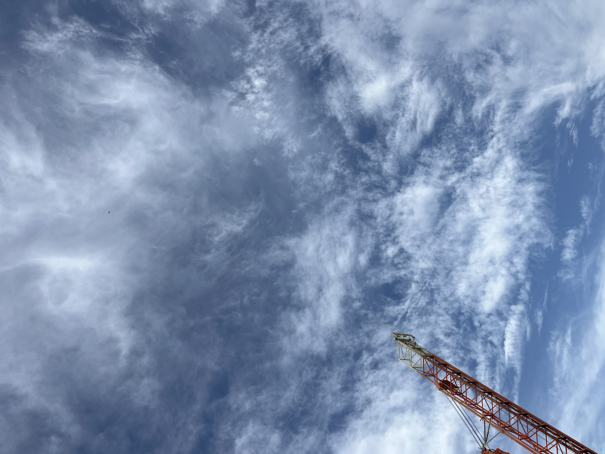 The image is a low-angle shot of a construction crane against a backdrop of blue sky partially covered by striated clouds. The crane is positioned in the lower right of the frame, and its lattice boom extends diagonally upward. The boom is painted red and white with a yellowish tip. The sky shows varying shades of blue with patches of white clouds streaking across the frame from left to right. A small, dark spot resembling a bird can be seen in the upper left quadrant of the sky. The setting is an urban environment in the city of Unknown, Unknown.