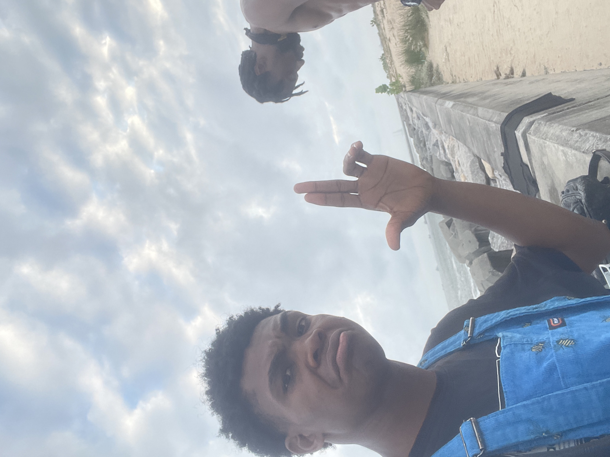 Two young Black males are on a sandy shore with a rocky embankment under a cloudy sky. The male in the foreground, wearing blue overalls over a black t-shirt, is looking at the camera with a displeased expression and has his left hand raised, fingers bent to form a gesture. The male in the background, shirtless, is looking down towards the rocky embankment. The location is Beecroft Point, Nigeria.