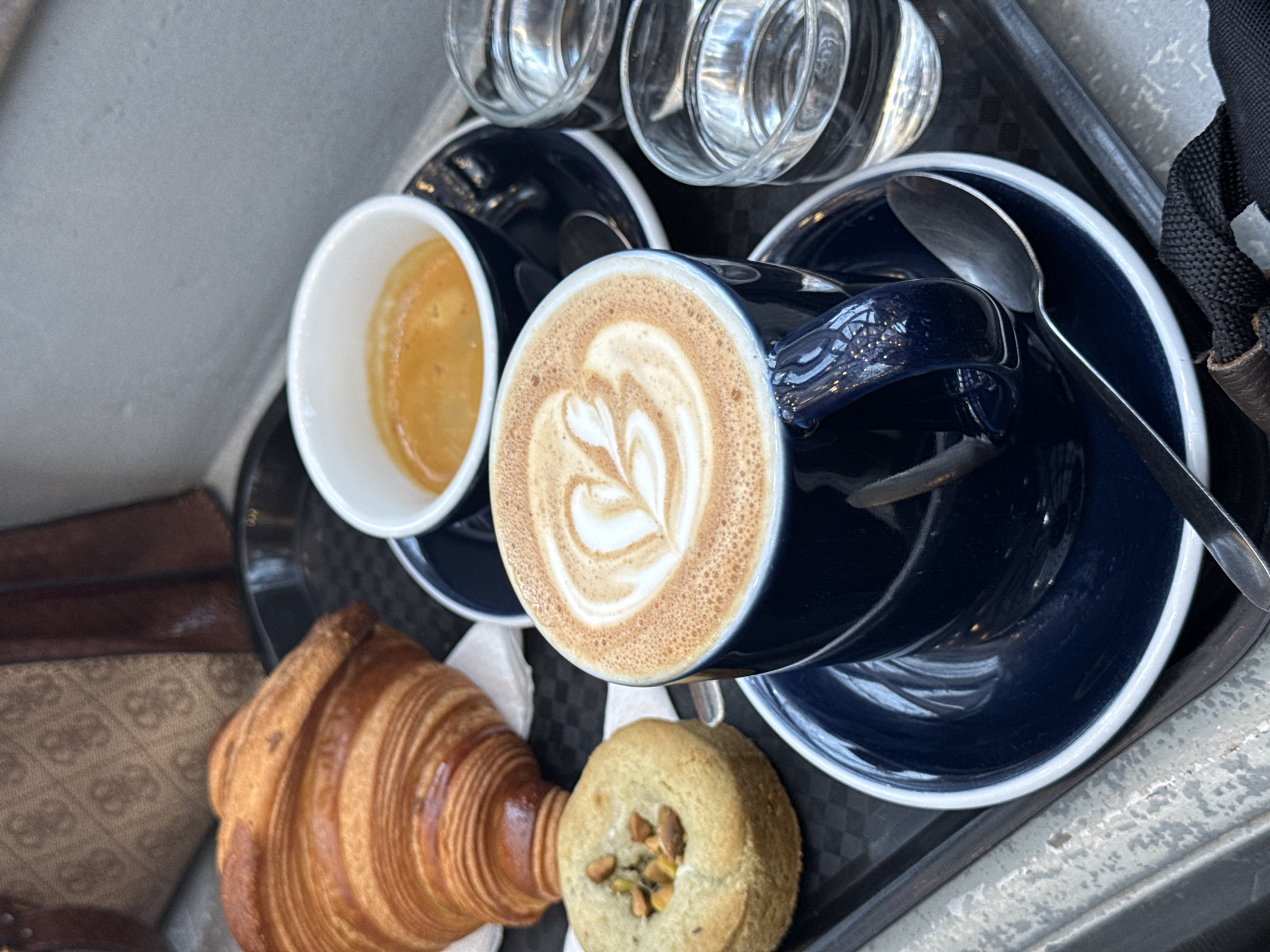 A high-angle, close-up photograph depicts a coffee and pastry service arranged on a textured black serving tray within an indoor establishment in Strasbourg, France. The tray contains a golden-brown croissant resting on a white napkin, and a small, round, light-brown pastry topped with chopped green and brown nuts, consistent with pistachios. Two coffee preparations are present: a small white cup with a dark rim holds an espresso with a rich amber crema, while a larger dark blue cup with a handle features a latte adorned with white heart and leaf latte art on its frothy surface. Both coffee cups sit on matching dark blue saucers with white rims, each accompanied by a small spoon. Two clear glasses, each containing approximately half a glass of water, are positioned to the right of the latte. In the upper left background, a portion of a light brown handbag with a repeating geometric pattern and dark trim is visible. The lower foreground displays a black fabric strap and a dark brown material. The background behind the tray is a plain, light grey wall or vertical surface.