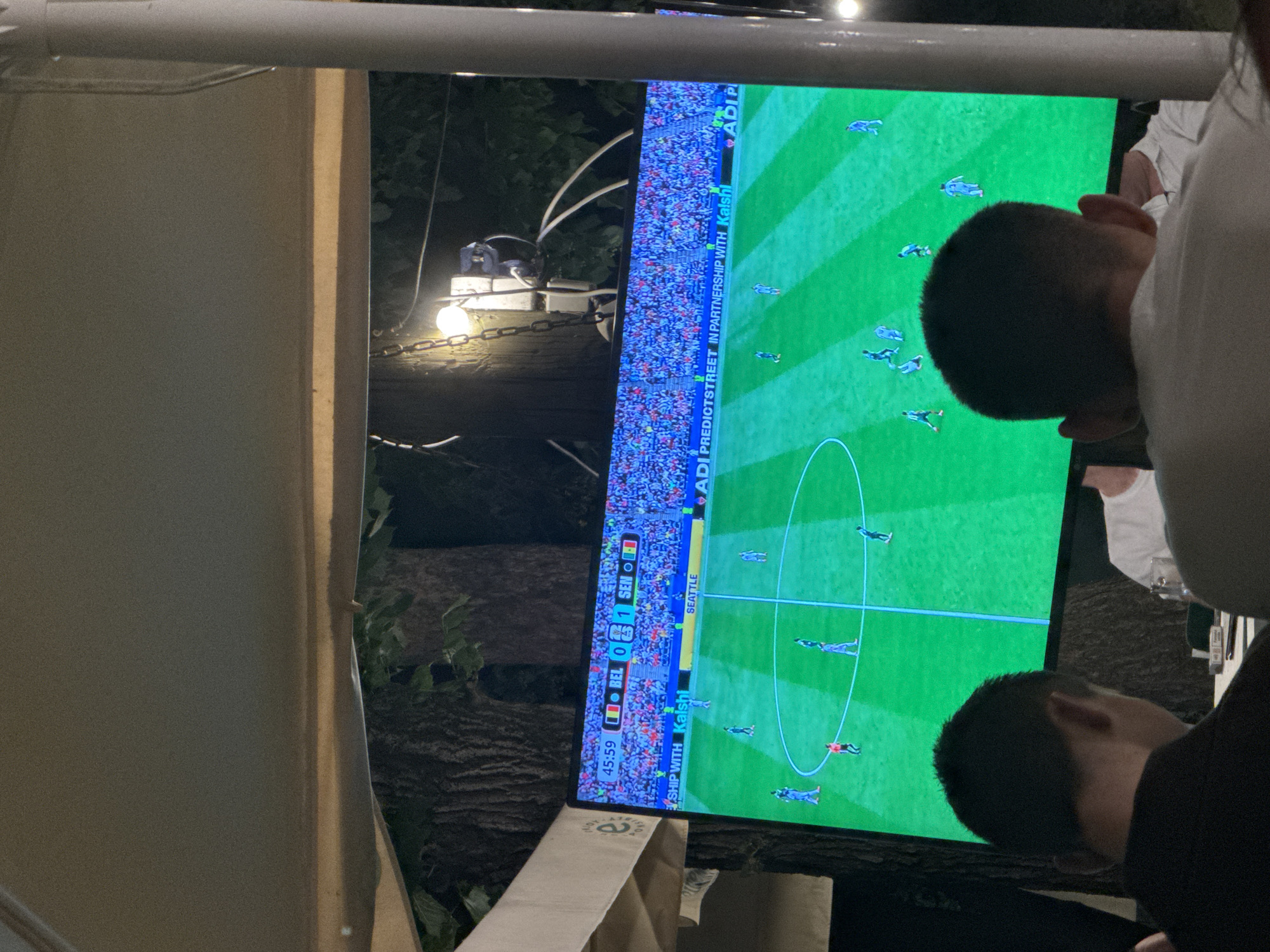The image depicts a scene in Suceava, Romania, where individuals are gathered to watch a soccer match on a large outdoor screen. The screen displays a game with the score showing Belgium (BEL) with 0 and Senegal (SEN) with 1. The time displayed is 45:59. The soccer field is vividly green, with players in blue uniforms on the screen. In the foreground, two men are visible from behind, engrossed in watching the match. One is wearing a black shirt, and the other, further back, is wearing a white shirt. The setting appears to be an outdoor seating area, possibly a restaurant or bar, with a canopy overhead and foliage in the background, suggesting an evening or nighttime event. A lamp hangs from a chain, illuminating the area. The overall atmosphere suggests a shared viewing experience of a sporting event.