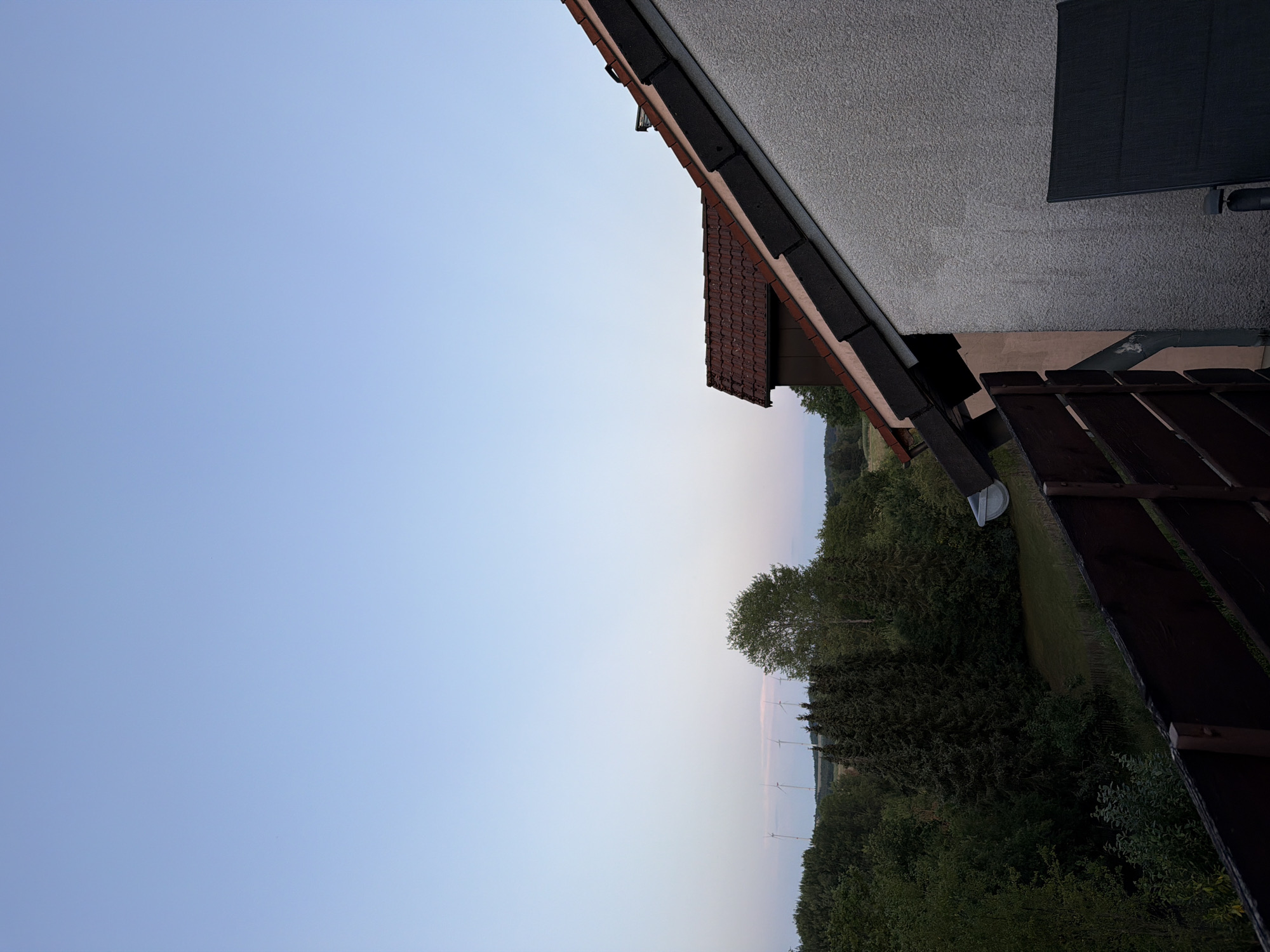 The image depicts a twilight view from an elevated position, likely a balcony or window, overlooking a rural landscape near Creußen, Germany. The foreground is dominated by a dark wooden railing. Beyond the railing, a lush green expanse of trees and a manicured lawn stretches towards the horizon. Several wind turbines are visible in the middle distance, silhouetted against the pale blue and pink hues of the fading sky. The sky itself transitions from a clear, light blue at the top of the frame to softer pastel shades near the horizon. On the right side of the image, the textured, light-colored wall of a building and its red-tiled roof are prominent, angled diagonally across the frame. A dark outdoor chair is partially visible in the lower right corner. The overall scene is peaceful and serene, characteristic of a rural setting at dusk.