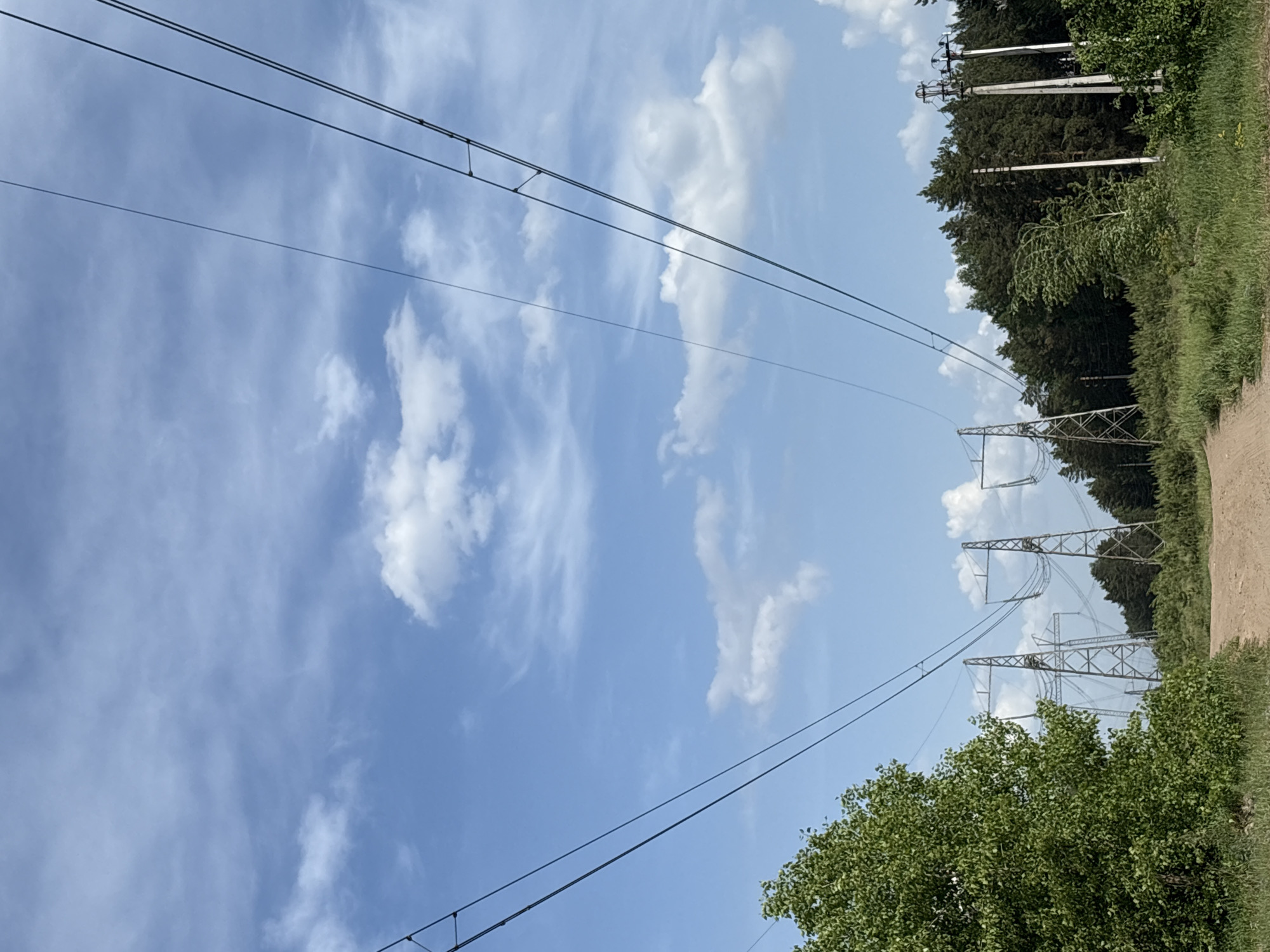 The image shows a view of the sky and power lines in Noviy, Russia. The sky is blue with scattered white clouds. Three sets of power lines stretch across the frame, connected to metal towers. On the right side of the frame, there's a dirt road alongside trees and vegetation. Some white posts, potentially for smaller power lines or utility poles, are visible among the trees. The scene appears to be taken from a rural or semi-rural area with power infrastructure running along a road or pathway near a wooded area.