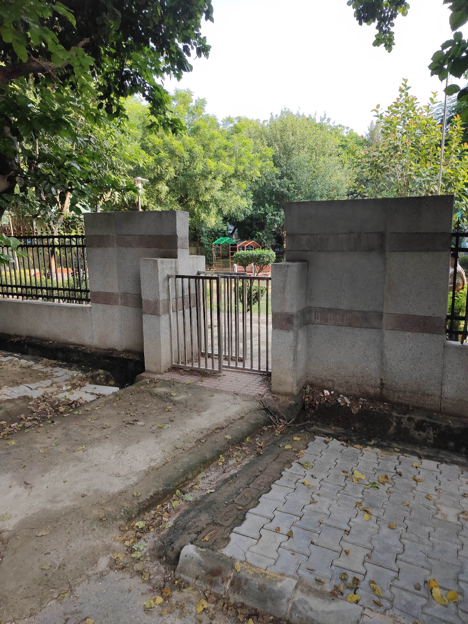 The image depicts a concrete gate structure in a park in Gurgaon, India. The gate is made of grey concrete blocks with horizontal bands of darker stone. A metal gate with vertical bars stands open. To the left of the gate is a black metal fence, and beyond that, trees and foliage. The ground in front of the gate is a mix of concrete and interlocking pavers, with fallen leaves scattered across both surfaces. In the background, more trees and a glimpse of playground equipment suggest a recreational area within the park. The lighting indicates it is daytime.