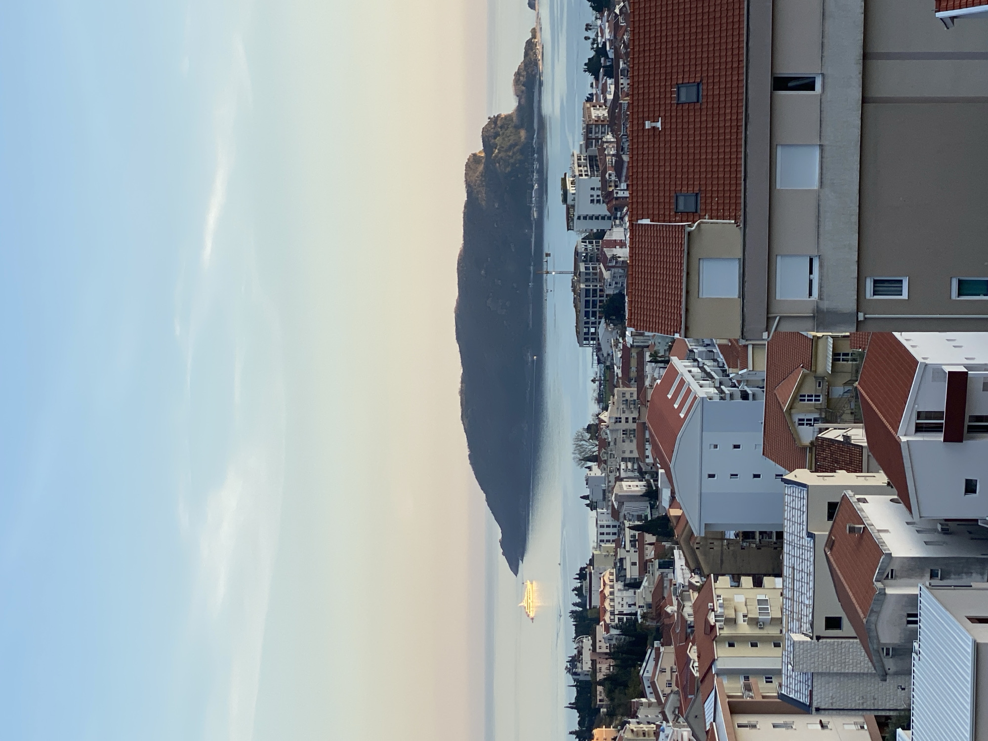 The image shows a cityscape with a body of water and a prominent land formation. The city features buildings with red tile roofs and light-colored facades. The water reflects sunlight, creating a bright streak. The land formation is dark and appears mountainous. The sky is pale blue. The location is likely Budva, Montenegro, based on the architectural style and coastal landscape.
