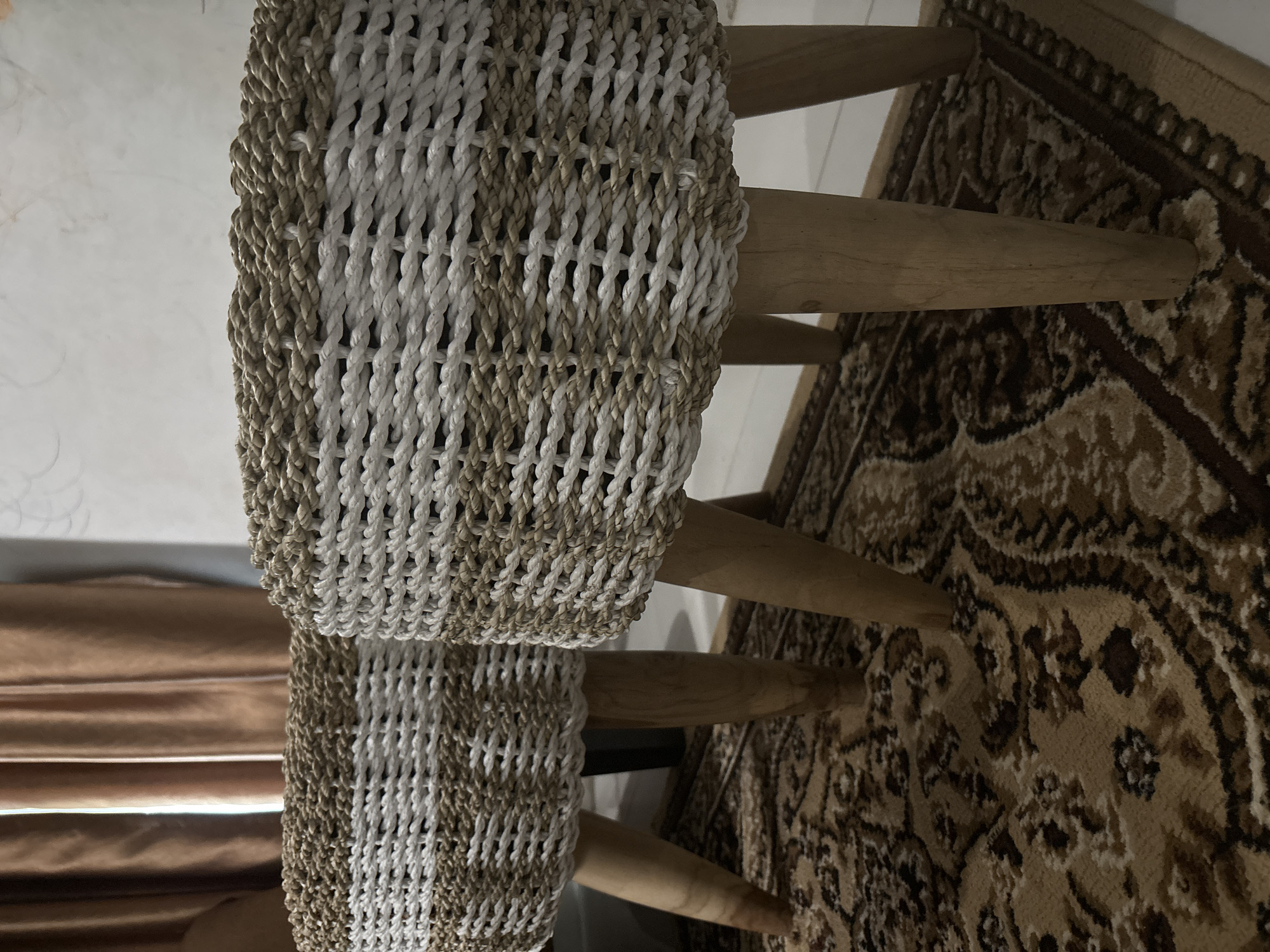 Two woven ottomans with wooden legs rest on a patterned rug. The ottomans are cylindrical with a white and tan woven pattern. The rug features a dark brown and tan paisley design. In the background, a brown curtain is visible. This scene is located in Geulanggang Teungoh, Indonesia.