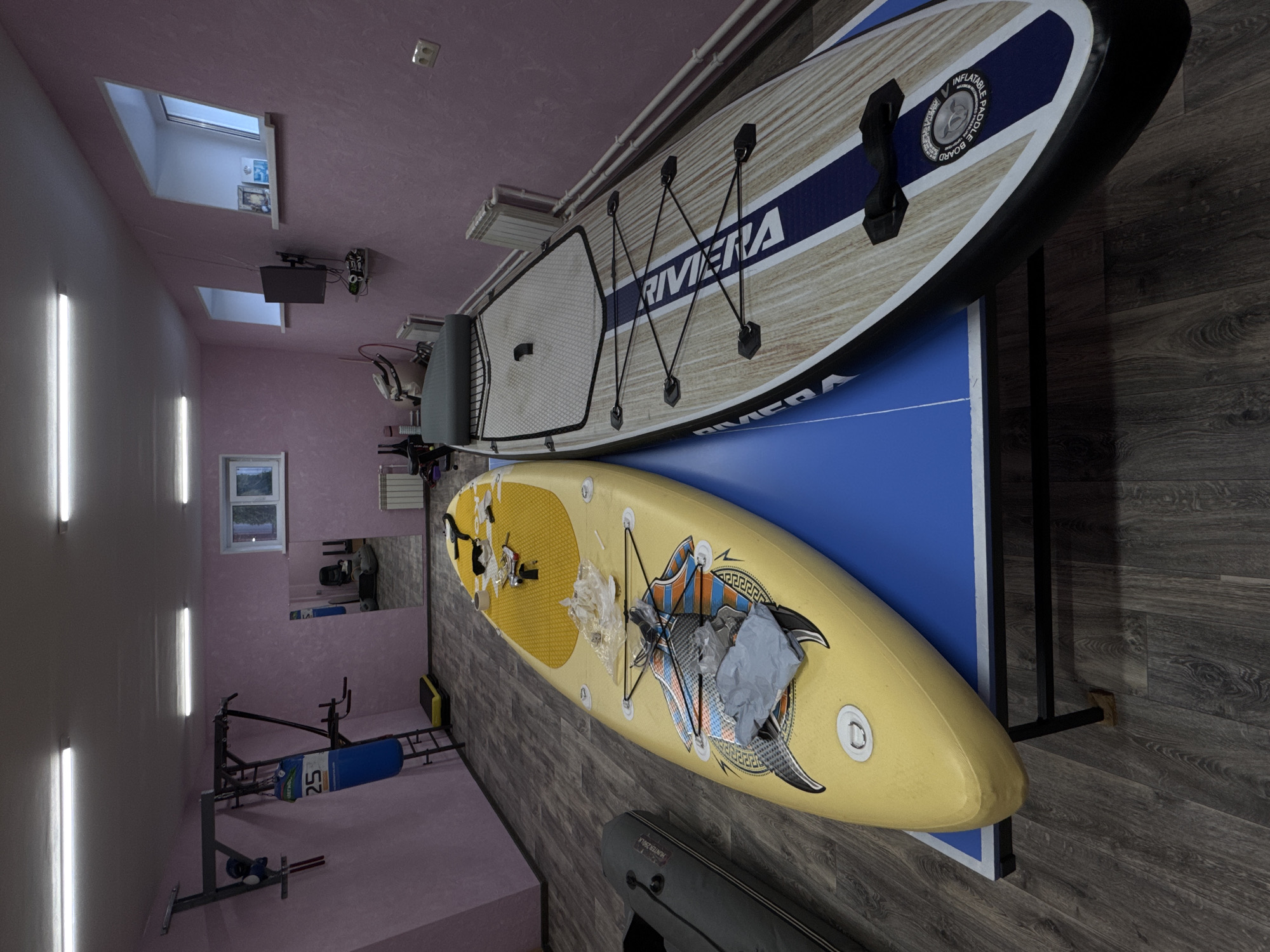 The image shows an indoor space, possibly a garage or recreational room, in Прикамский, Russia. The walls and ceiling are a light purple color, and the floor is a dark wood-grain laminate. Fluorescent lights are installed on the walls.
Two paddleboards are propped against a ping pong table. The top paddleboard has a wood grain pattern and a blue stripe with the word "RIVIERA" on it. The second paddleboard is yellow with decorative graphics.
Exercise equipment, including a weight bench and weights, is present in the room, indicating it is used for fitness activities. The window provides natural light.