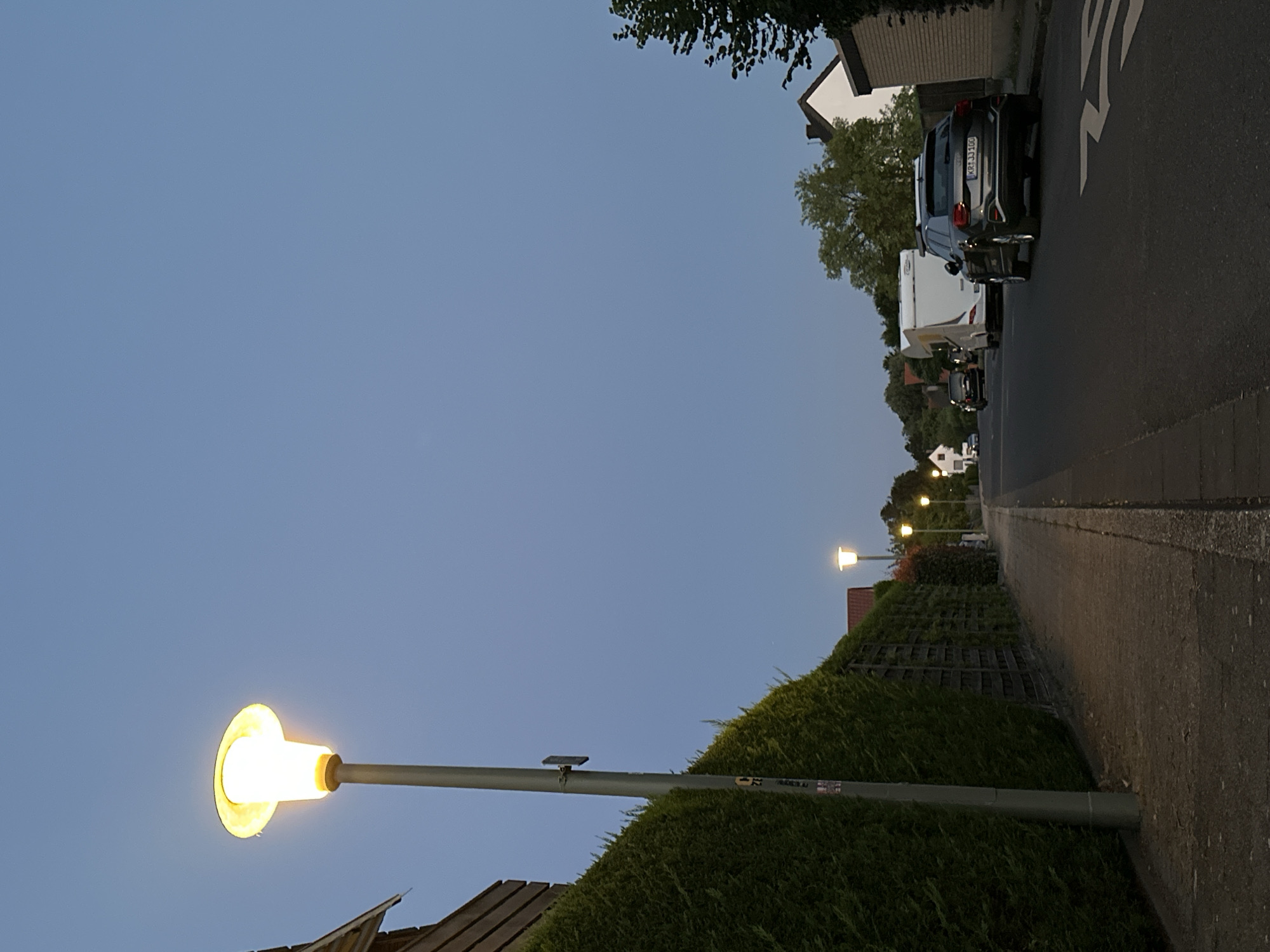 The image captures a street scene in Krefeld, Germany, during twilight. A tall lamppost with its light illuminated stands on the left side of the image, casting a warm glow against the dusky blue sky. Adjacent to the lamppost is a dense green hedge that runs along a sidewalk. Further down the street, several houses with gabled roofs are visible, interspersed with trees. A line of streetlights, also illuminated, stretches into the distance along the right side of the road. Parked on the right side of the asphalt road are a dark-colored SUV and a white caravan. The license plate on the SUV reads "KR 1JJ 100". The overall atmosphere is quiet and residential, with the dimming natural light and artificial streetlights creating a serene evening ambiance.