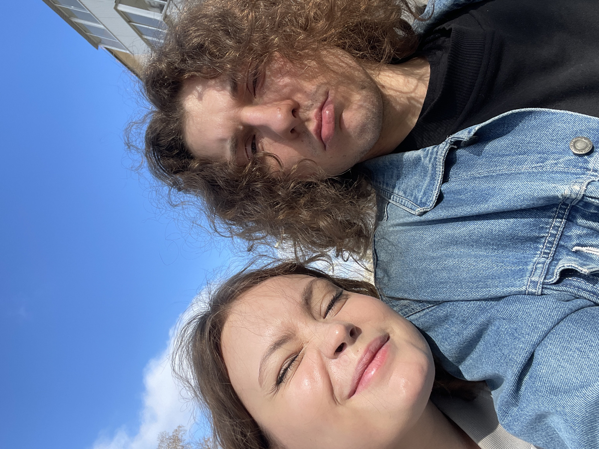 Two individuals are pictured from a low-angle perspective. The person at the top has curly brown hair, closed eyes, and is wearing a black shirt. The person at the bottom has light brown hair, closed eyes, and a denim jacket over what appears to be a light-colored shirt. The backdrop is a bright blue sky with sparse clouds. The angle suggests they are lying down. The presence of a building in the background indicates an urban environment, potentially Yekaterinburg.