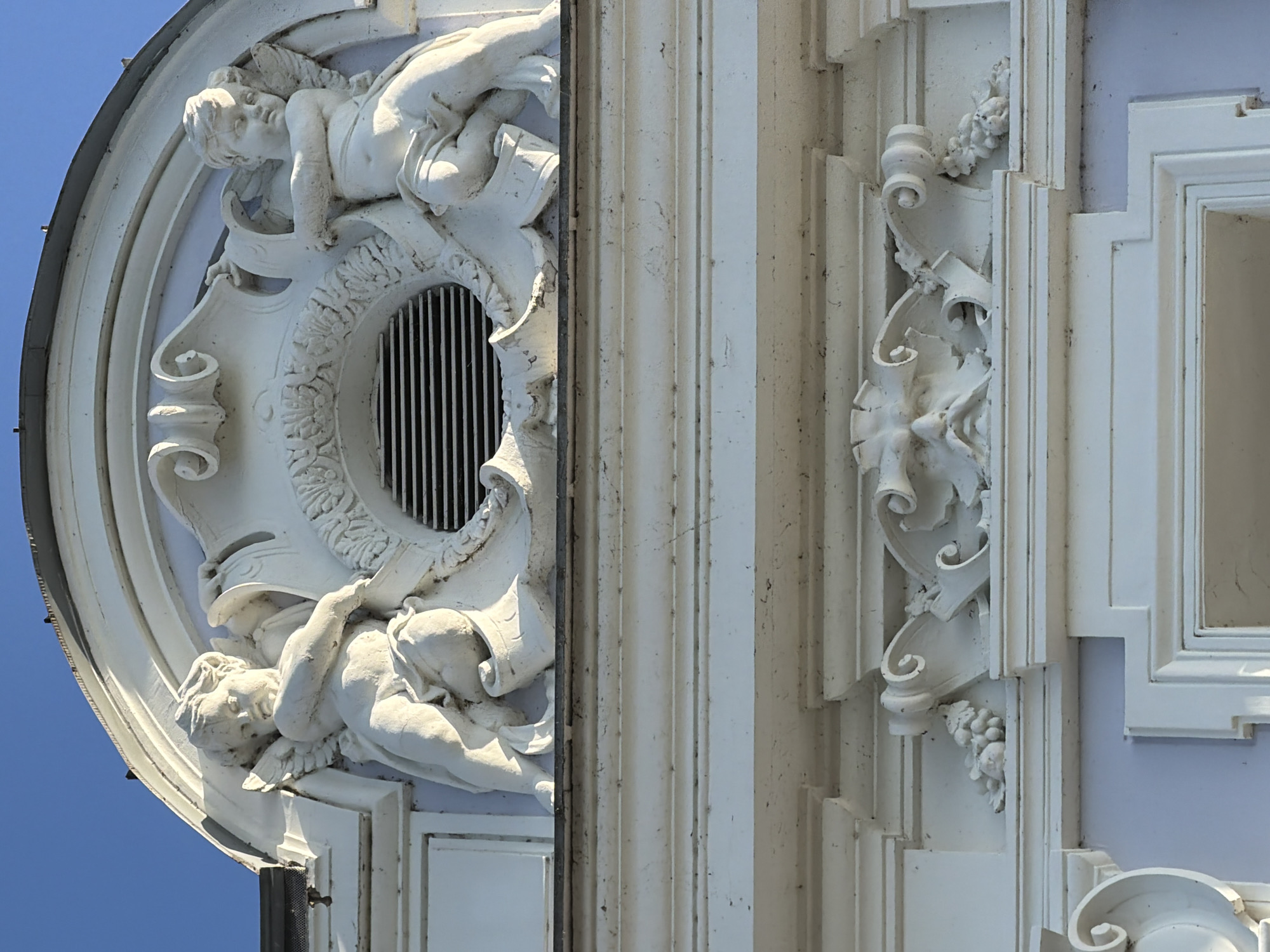 The image shows a detailed architectural facade with white stucco ornamentation against a clear blue sky. The primary focus is on a large, decorative element featuring two cherubic figures with wings, one on each side, positioned above a circular vent. These figures appear to be made of plaster or stone, with delicate detailing. Between the cherubs, a wreath-like design surrounds the vent. Below this central element, another decorative band features a grotesque mask with a mustache and beard, flanked by clusters of grapes. The architectural style suggests a historical building, possibly a public institution or a grand residence. The image is a close-up shot, highlighting the intricate craftsmanship of the facade. The location is identified as Nizhny Novgorod, Russia.