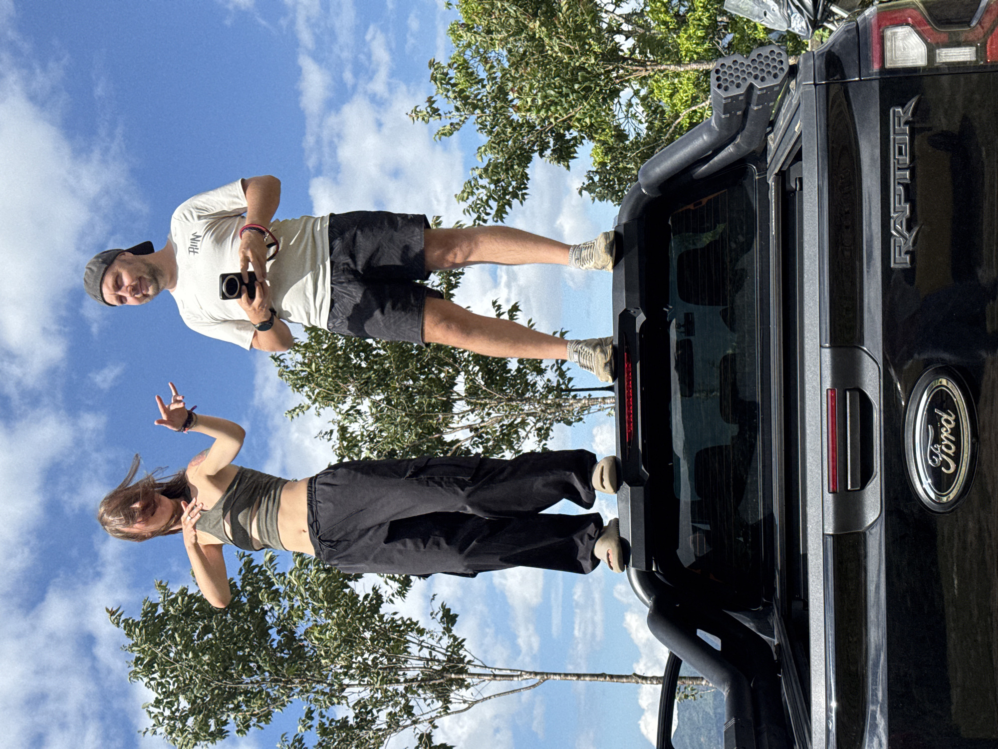 In Tu Le, Vietnam, two individuals are standing on the roll bar of a black Ford Raptor pickup truck, positioned against a backdrop of a bright blue sky with scattered white cumulus clouds and green trees. The individual on the left, a female with light brown hair swept across her face, is wearing an olive-green halter-neck crop top with a midriff cut-out and loose-fitting black cargo pants with elasticated ankles. She has light-colored slip-on sandals on her feet. Her right hand (viewer's left) displays a "rock on" gesture with extended index and pinky fingers, while her left hand is bent near her chest. A tattoo is visible on her right shoulder area. The individual on the right, a male, wears a dark grey baseball cap backward, a light beige t-shirt with a dark graphic, and dark patterned athletic shorts. He has light-colored athletic shoes on his feet. He holds a dark-colored smartphone vertically with both hands and wears a dark watch on his left wrist (viewer's right) and a bracelet on his right wrist (viewer's left), gazing towards the left of the frame. The truck's tailgate displays the "Ford" logo and "RAPTOR" emblem. A red accent strip is visible above the tailgate handle. An accessory with multiple circular openings is mounted on the right side of the truck bed, near the rear. The background consists of verdant trees under the clear sky.