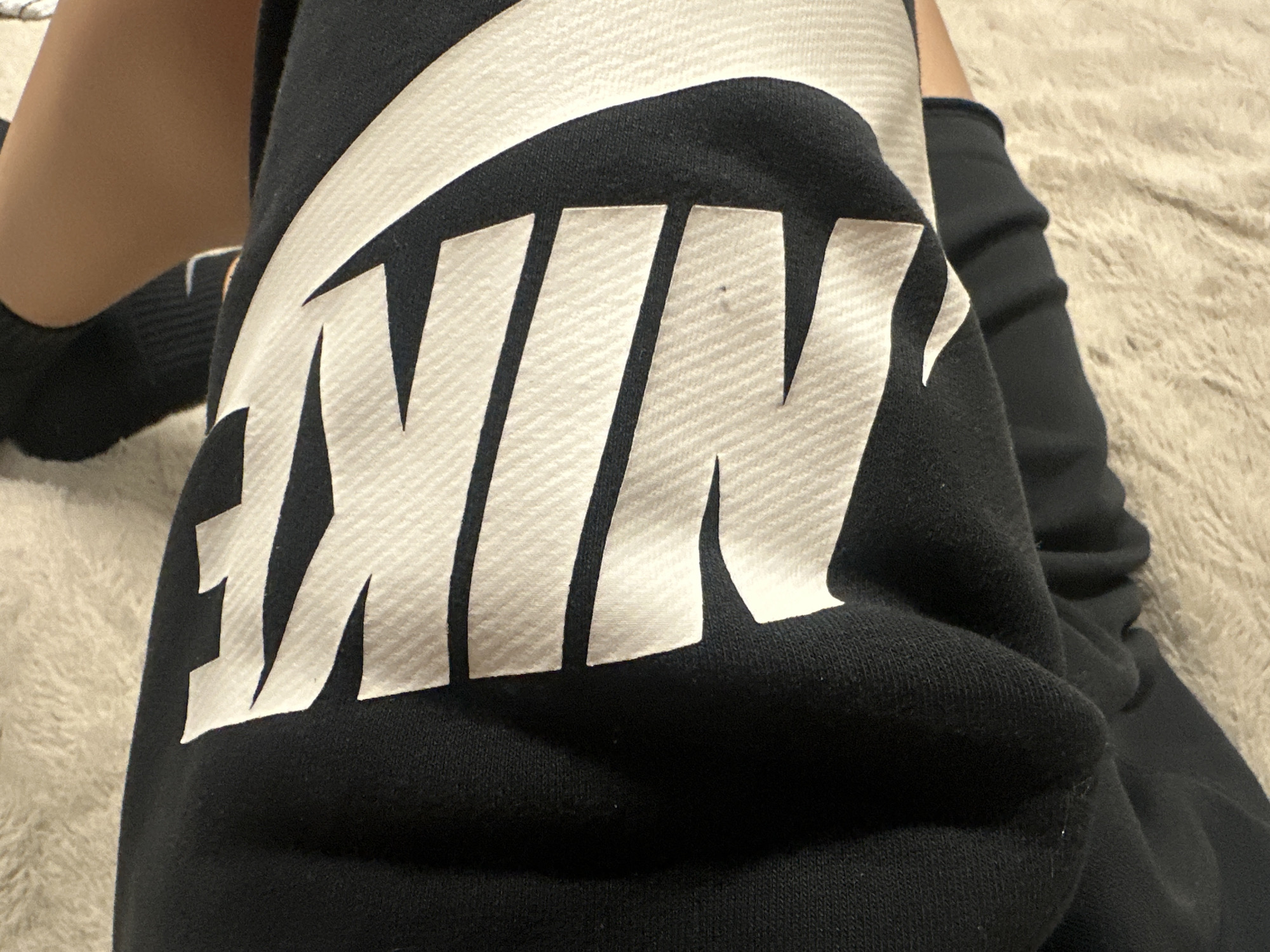 A close-up image shows a person resting, wearing black athletic wear featuring a large white "NIKE" logo. The logo, complete with its characteristic swoosh, is prominently displayed across the fabric, which appears to be a black cotton or fleece material, possibly part of sweatpants or a sweatshirt. Subtle textural patterns are visible within the white letters of the logo and on the black fabric. A small, dark speck is visible on the letter "I." To the left of the garment, a segment of a bare human leg with light-to-medium skin tone is visible, ending in a foot and ankle covered by a plain black sock. The background consists of a light, neutral-colored, plush, and textured material, resembling a shag carpet or a soft blanket, suggesting an indoor setting.