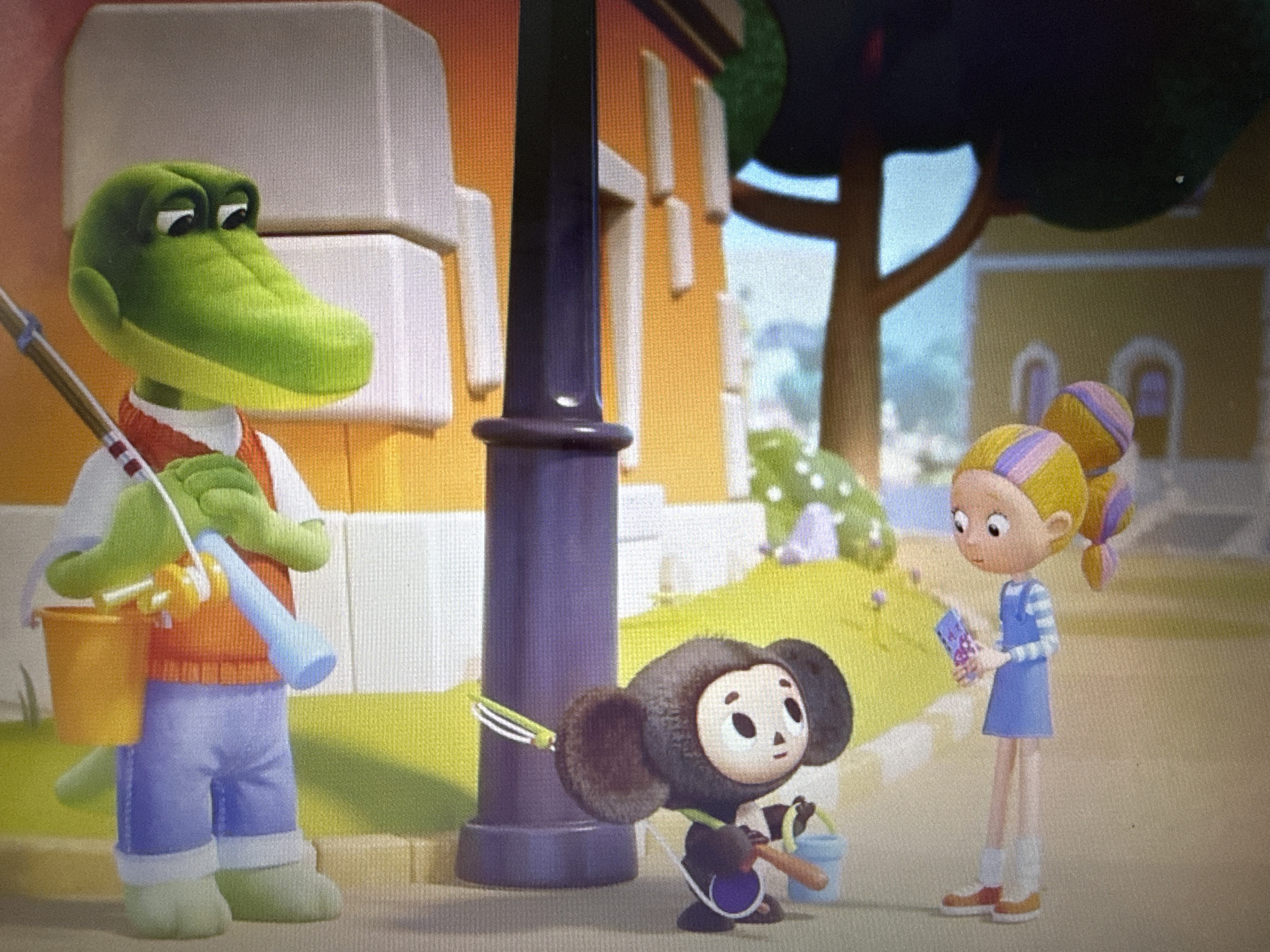 In a cartoonish outdoor scene set in Pushkino, Russia, three animated characters are depicted. On the left stands a green alligator wearing an orange vest over a white shirt and blue pants. He is holding a fishing rod and a yellow bucket. In the center, a small, dark-furred creature resembling a bear cub, possibly Cheburashka, is crouched down, holding a small blue bucket and a stick. To the right, a young girl with blonde hair tied in pigtails, wearing a blue dress over a striped long-sleeved shirt and white socks, is standing and holding a small purple object. The setting appears to be a sunny day in a suburban area, with buildings and trees in the background, suggesting a residential street or park. The overall mood is light and playful, as the characters seem to be engaged in a casual outdoor activity, possibly fishing or playing. No visible text is present in the image.