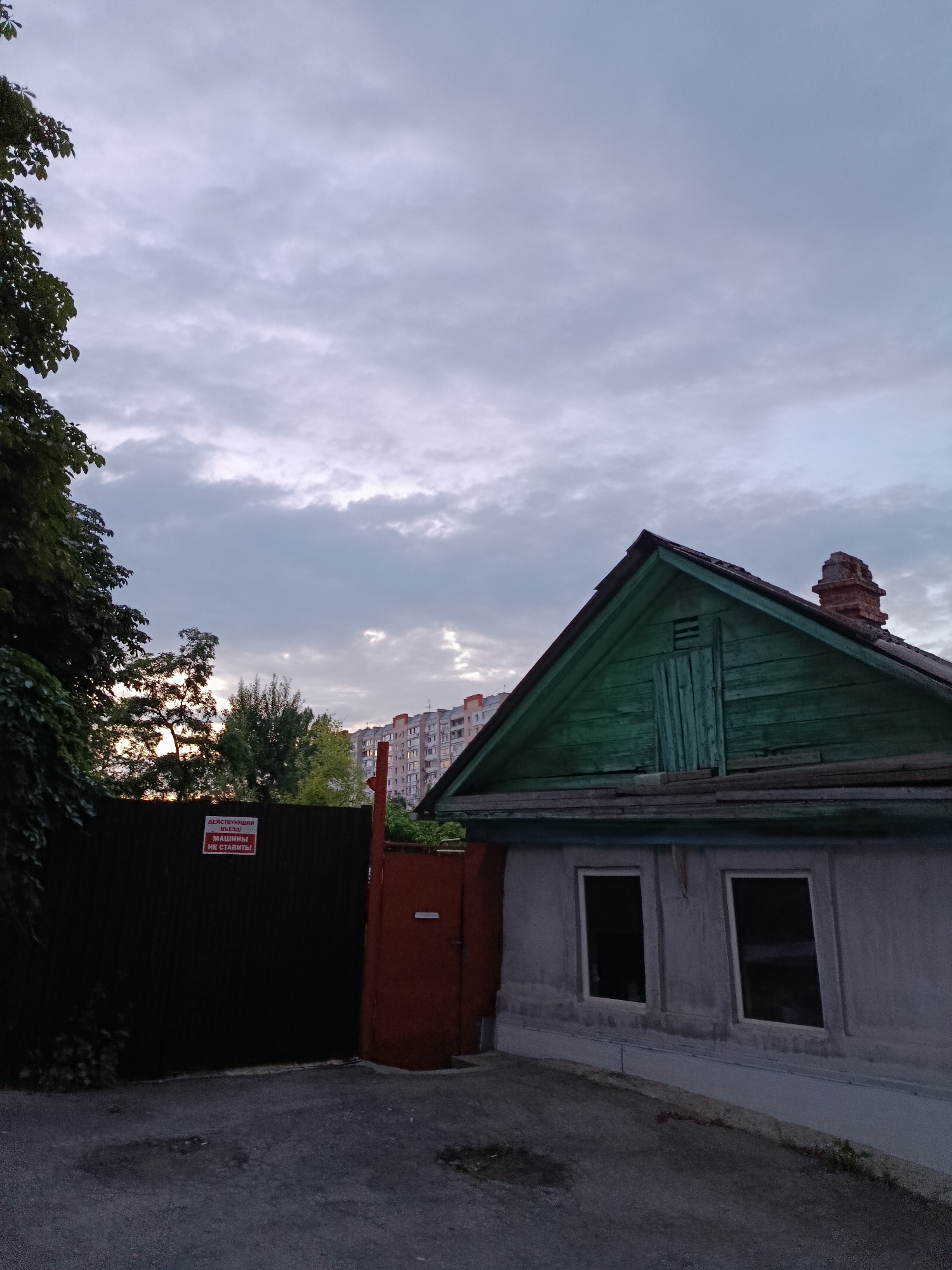 The image depicts a residential building with a green-painted wooden gable roof and a brick chimney, set against a cloudy sky. Adjacent to this building, on the left, is a dark metal gate with a red-painted door and a sign that reads "ДЕЙСТВУЮЩИЙ ВЪЕЗД! МАШИНЫ НЕ СТАВИТЬ!" which translates to "ACTIVE ENTRANCE! DO NOT PARK CARS!". Behind the gate, trees are visible, and further in the background, a multi-story apartment building can be seen. The ground in front of the gate and building is asphalted. The scene appears to be in an urban environment, likely in Saratov, Russia, given the Cyrillic text on the sign. No people are present in the image. The overall mood is somewhat somber, possibly due to the overcast sky.