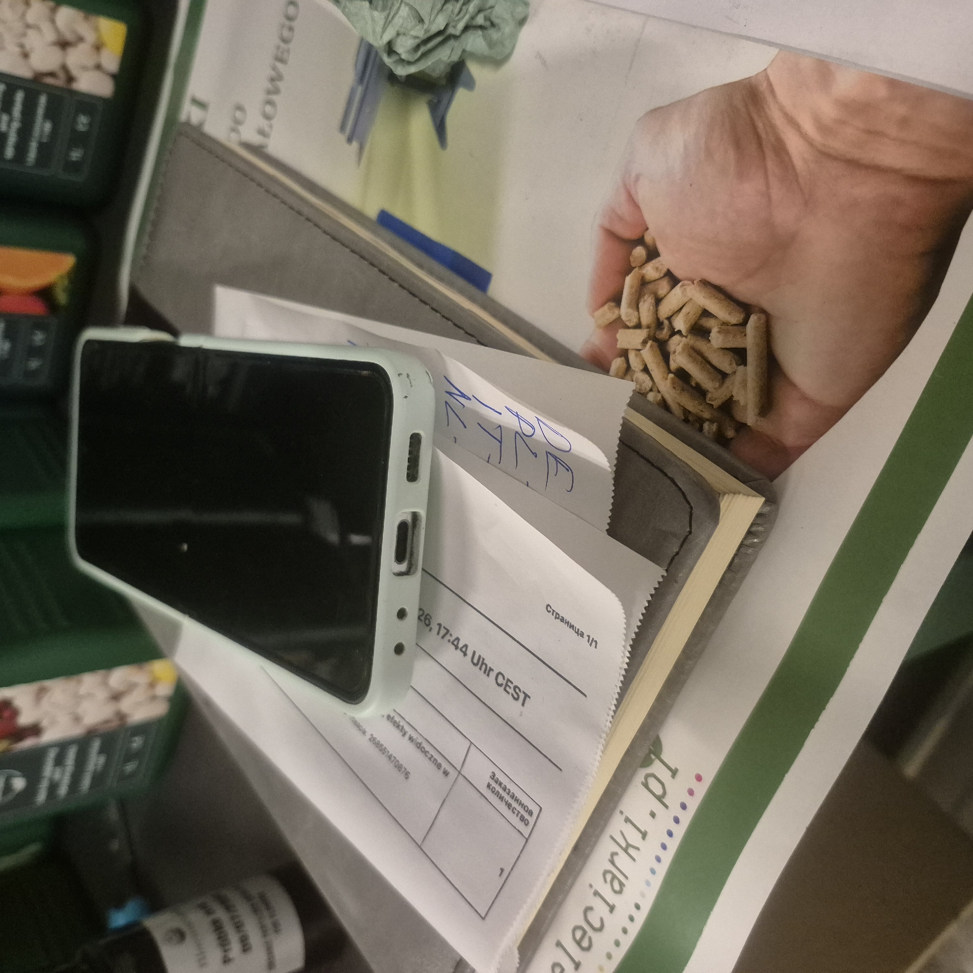 A smartphone with a light blue case rests on top of a notebook. The notebook is on top of a document with printed text in Polish. In the foreground, a hand holds a pile of light brown pellets. The background shows out-of-focus green containers. The document has a date stamp reading "26.17:44 UHR CEST". The text "eleciarki.pl" is visible on a green and white striped paper in the lower left corner of the image. The location is not specified in the image.