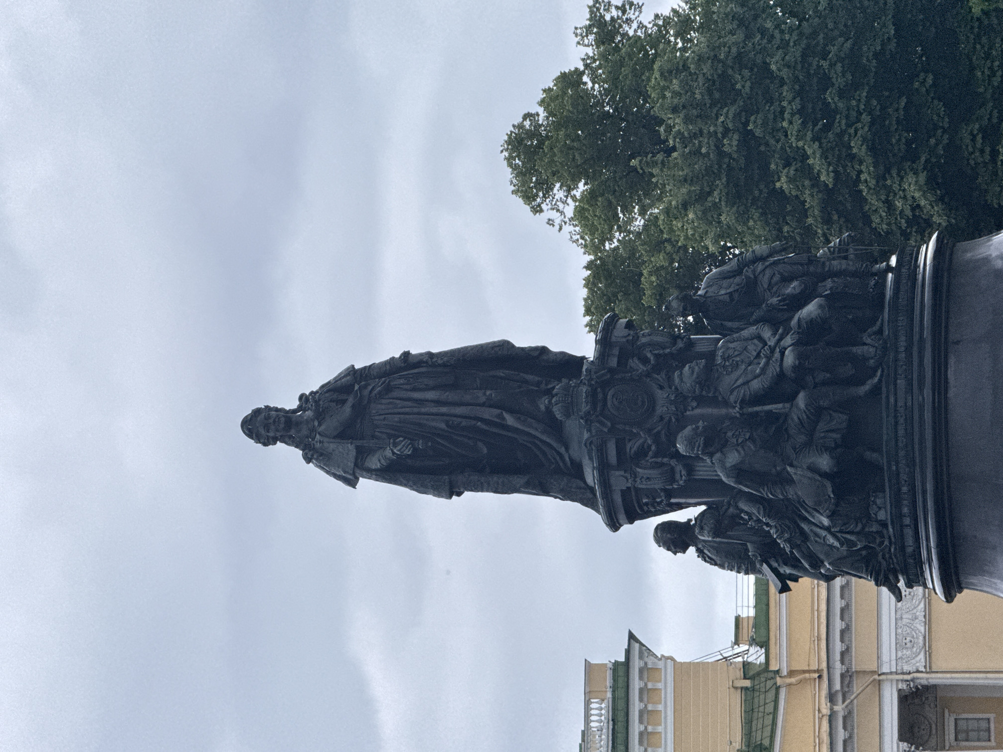 The image shows a monument in Saint Petersburg, Russia, likely the Monument to Catherine the Great in Nevsky Prospekt. The monument is a large bronze sculpture with a central figure of Catherine the Great standing on a pedestal. Below her, several smaller figures are depicted in various poses. The monument is dark, appearing to be made of bronze. In the background, a yellow building with white architectural details and green accents is visible, along with lush green trees under a cloudy, overcast sky. The overall atmosphere of the image is somber and majestic.