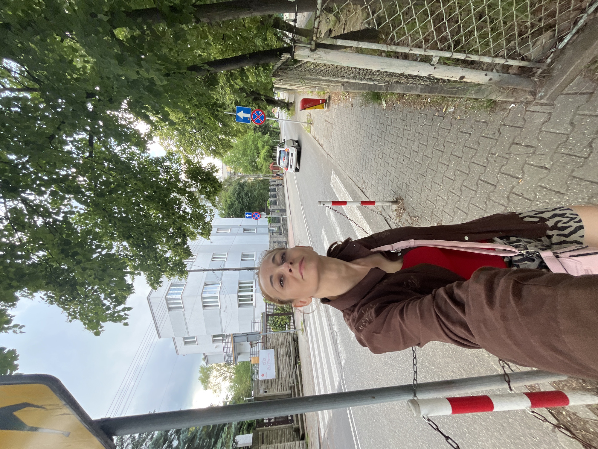 In this image, a young woman with blonde hair is taking a selfie in an outdoor urban setting. She is wearing a brown jacket over a red top and patterned shorts, with a pink purse slung over her shoulder. Her expression is neutral as she looks towards the camera.
The scene appears to be a street with a pedestrian crossing. The street is paved with interlocking bricks, and there's a sidewalk alongside it. Trees with lush green leaves line the street, providing shade. Across the street, a modern, multi-story building with white facades and many windows stands prominently. There are also traffic signs visible, including a blue sign with a white arrow and a red circle with a diagonal line, indicating "no entry" or a restricted zone. A white car is seen driving on the road.
The lighting suggests it is daytime, possibly late morning or afternoon, with sunlight filtering through the leaves. The sky is overcast, indicating a cloudy day. The presence of a chain and a red and white striped barrier suggests some form of demarcation or temporary closure on the sidewalk or street. The overall impression is of a casual day out in a European-style city. There is some visible text on a white sign on the building on the right, but it is too blurry to read.