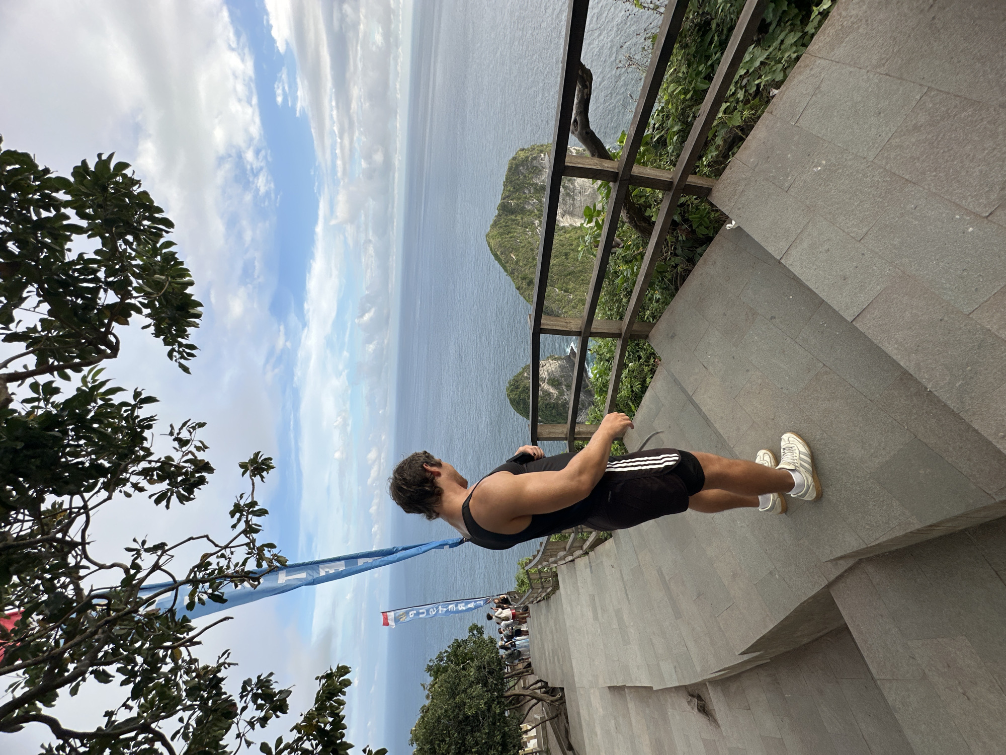 A man in a black tank top and black shorts with white stripes stands on a stone staircase. He faces away from the camera and toward the ocean and a nearby cliff covered in vegetation. A wooden railing with vegetation surrounds part of the stairs. In the background, flags are blowing in the wind. Trees are on the left side of the image, and the sky is filled with clouds. The location appears to be in Nusa Penida, Indonesia.
